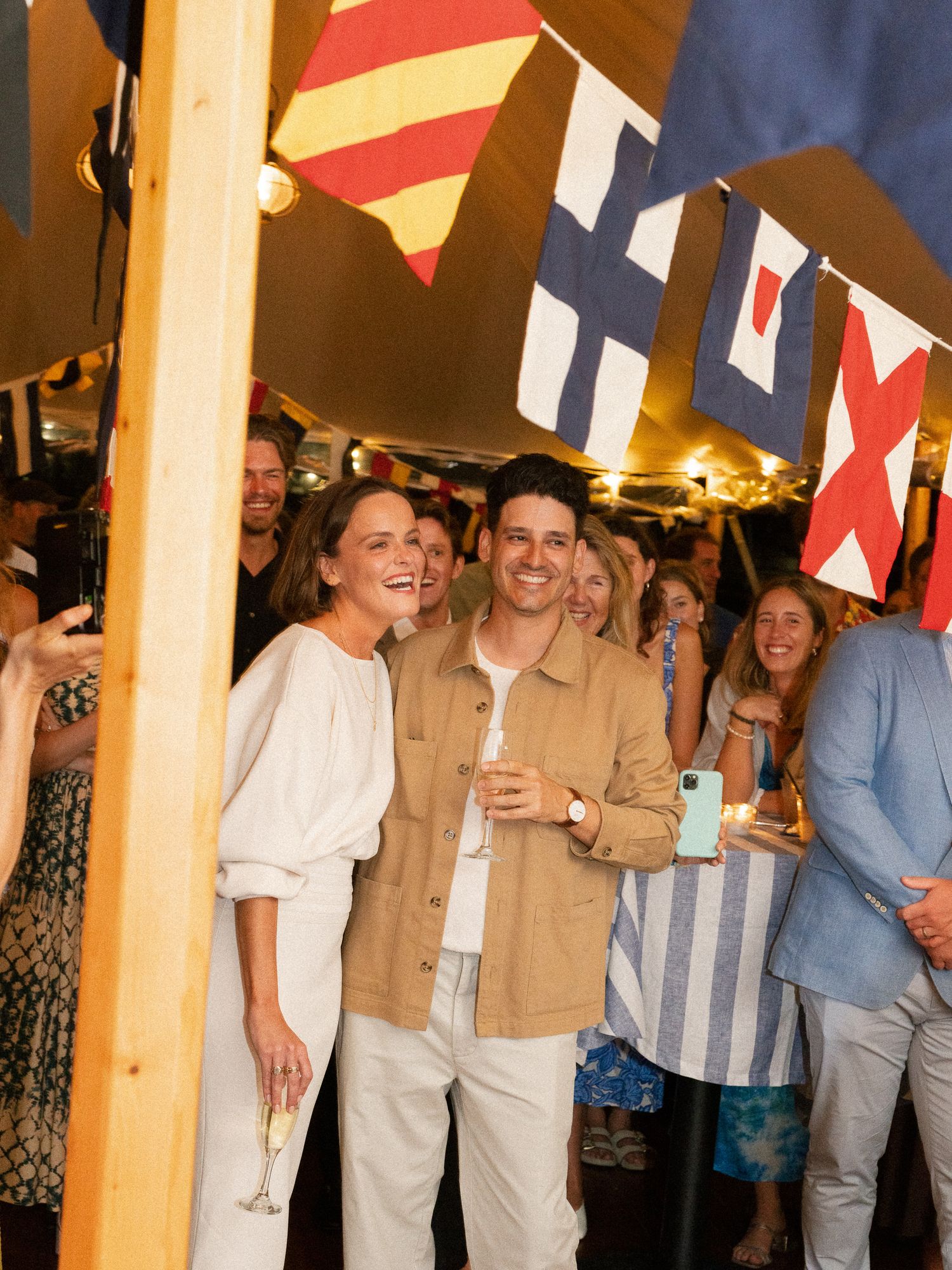 Couple smiling under colorful nautical signal flags at an outdoor tented wedding welcome party.