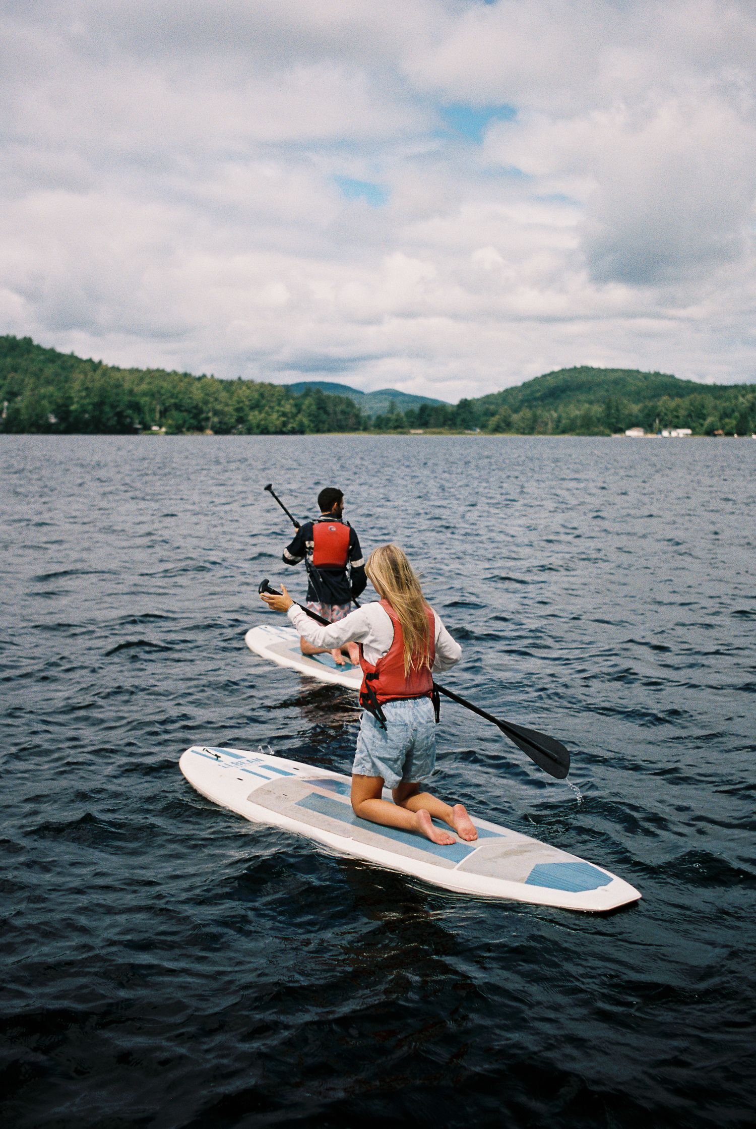 Two people in life jackets paddleboarding on a lake with green mountains in the distance.