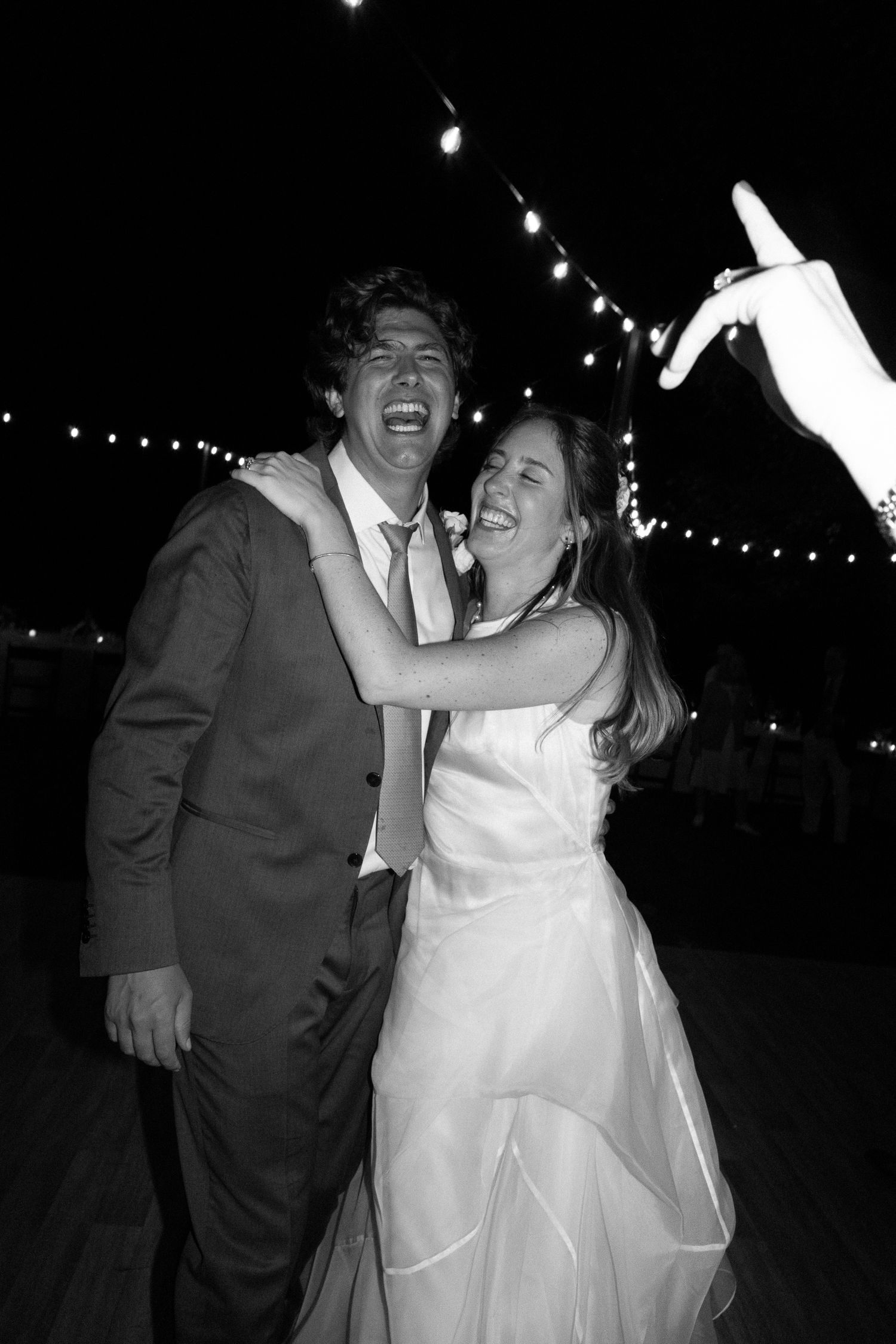 Joyful bride and groom laughing together on dance floor under string lights at outdoor wedding reception.