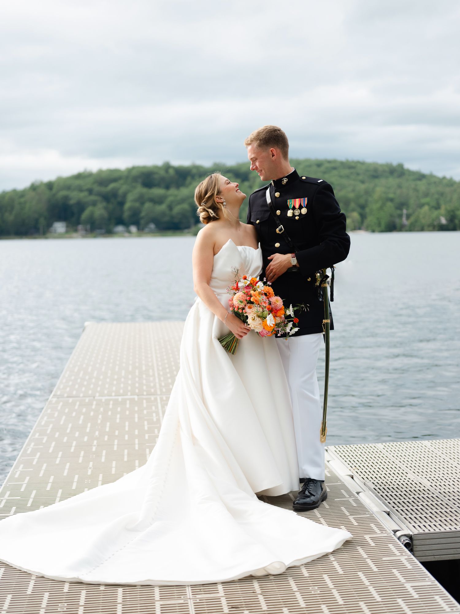 Military groom in dress uniform and bride in strapless gown pose on lake dock with colorful bouquet.
