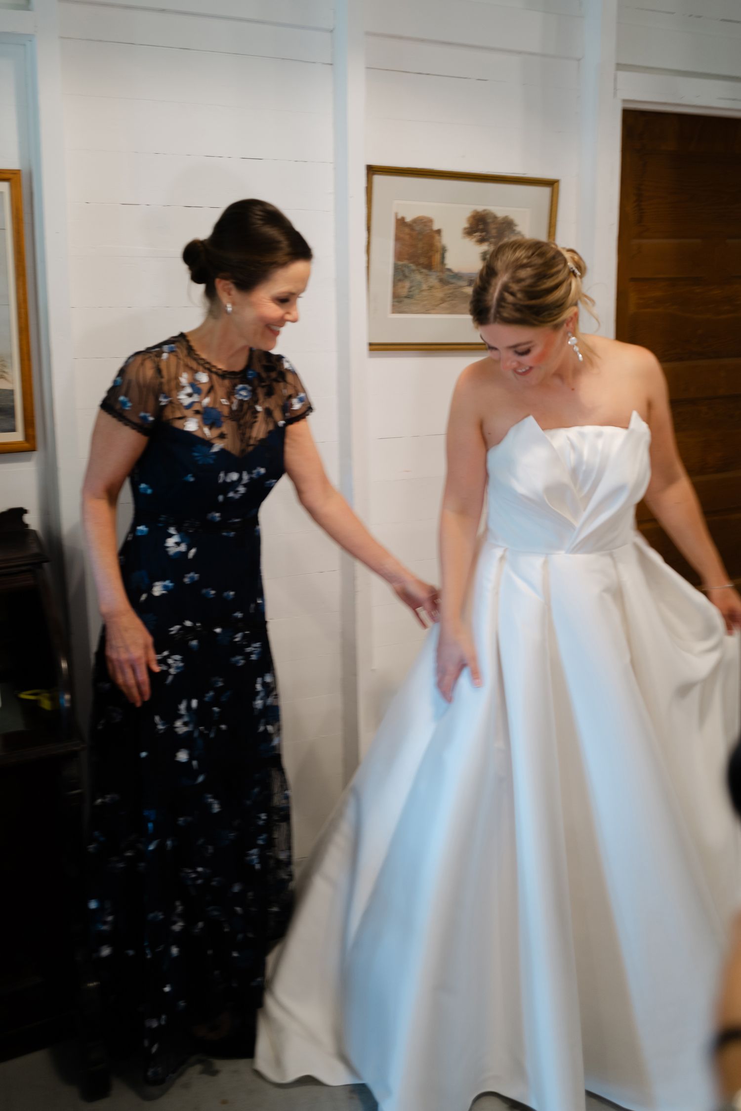 Bride in strapless white ball gown laughing with woman in navy floral dress indoors.