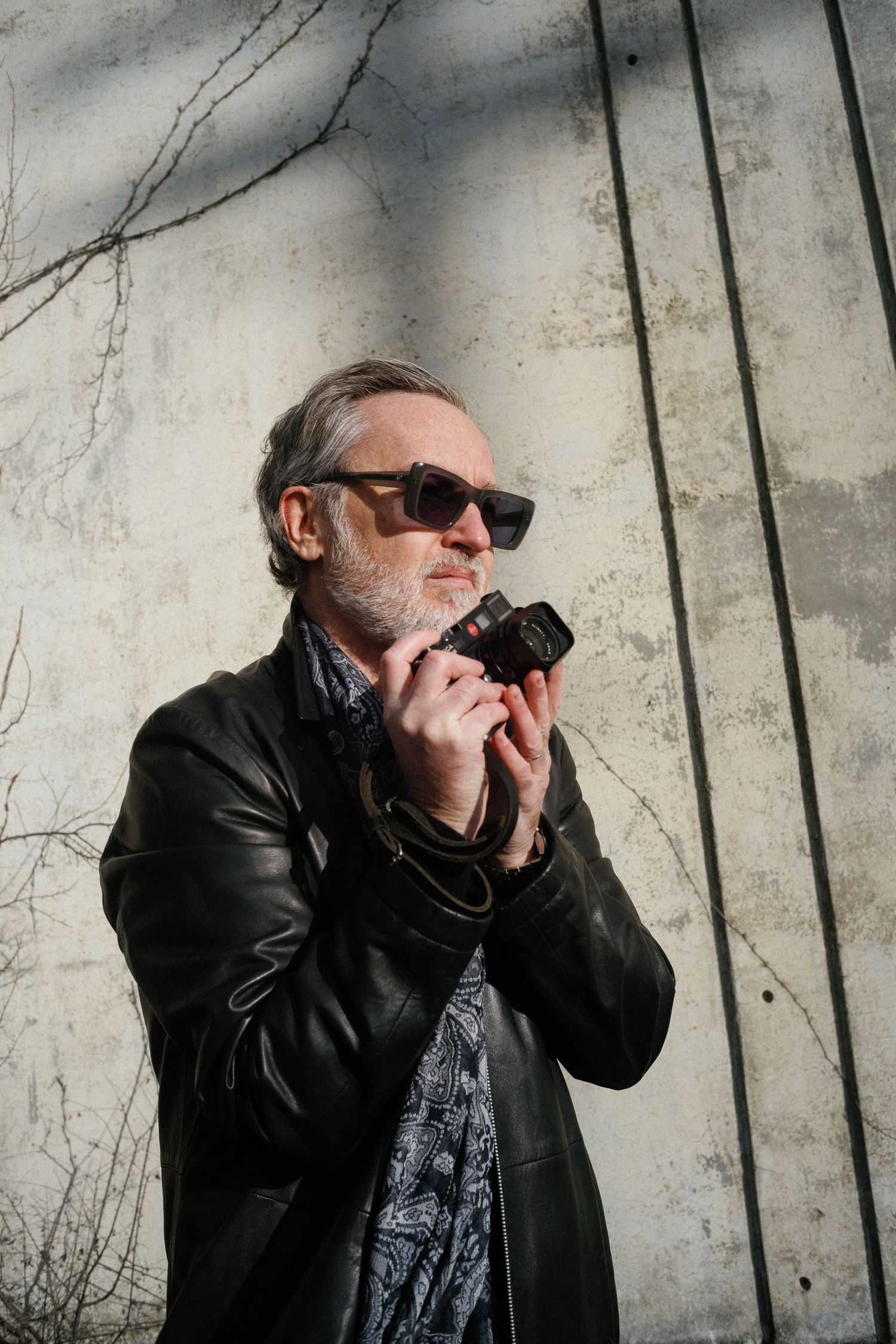 Middle-aged man with gray hair and beard wearing sunglasses and leather jacket holding vintage camera outdoors.