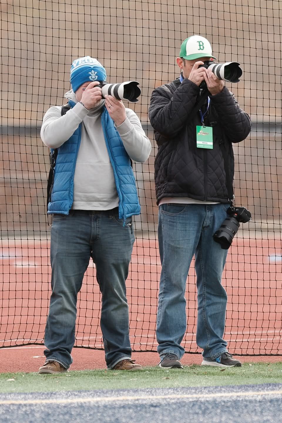 Two photographers with telephoto lenses shoot at a sporting event near a net on a track field.