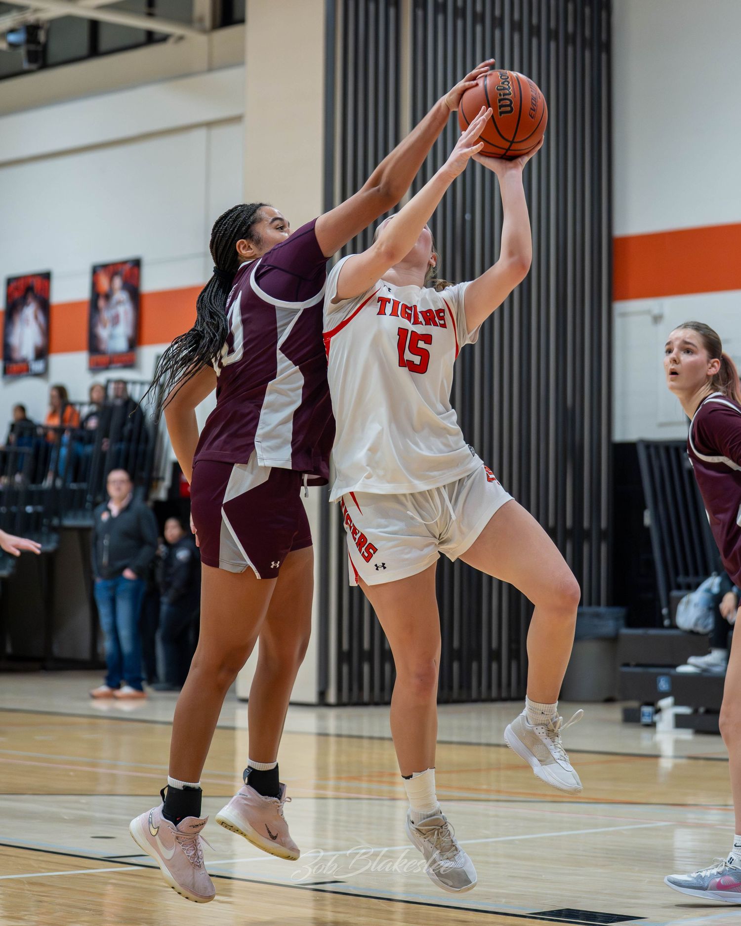 Female basketball player #15 in white attempts a layup while defender in maroon tries to block the shot.