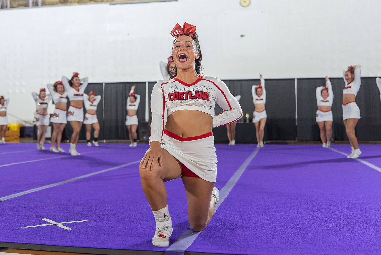 Cortland cheerleader performs energetic routine on purple mat at cheerleading competition.