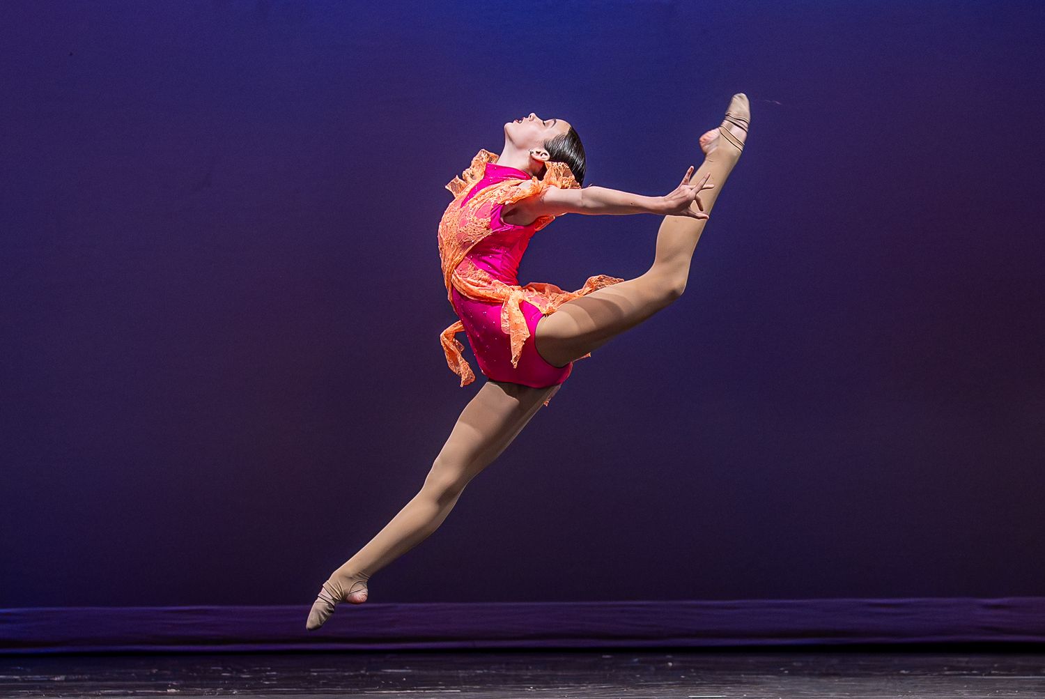 A dancer in a vibrant pink costume leaps gracefully mid-air on a purple-lit stage.