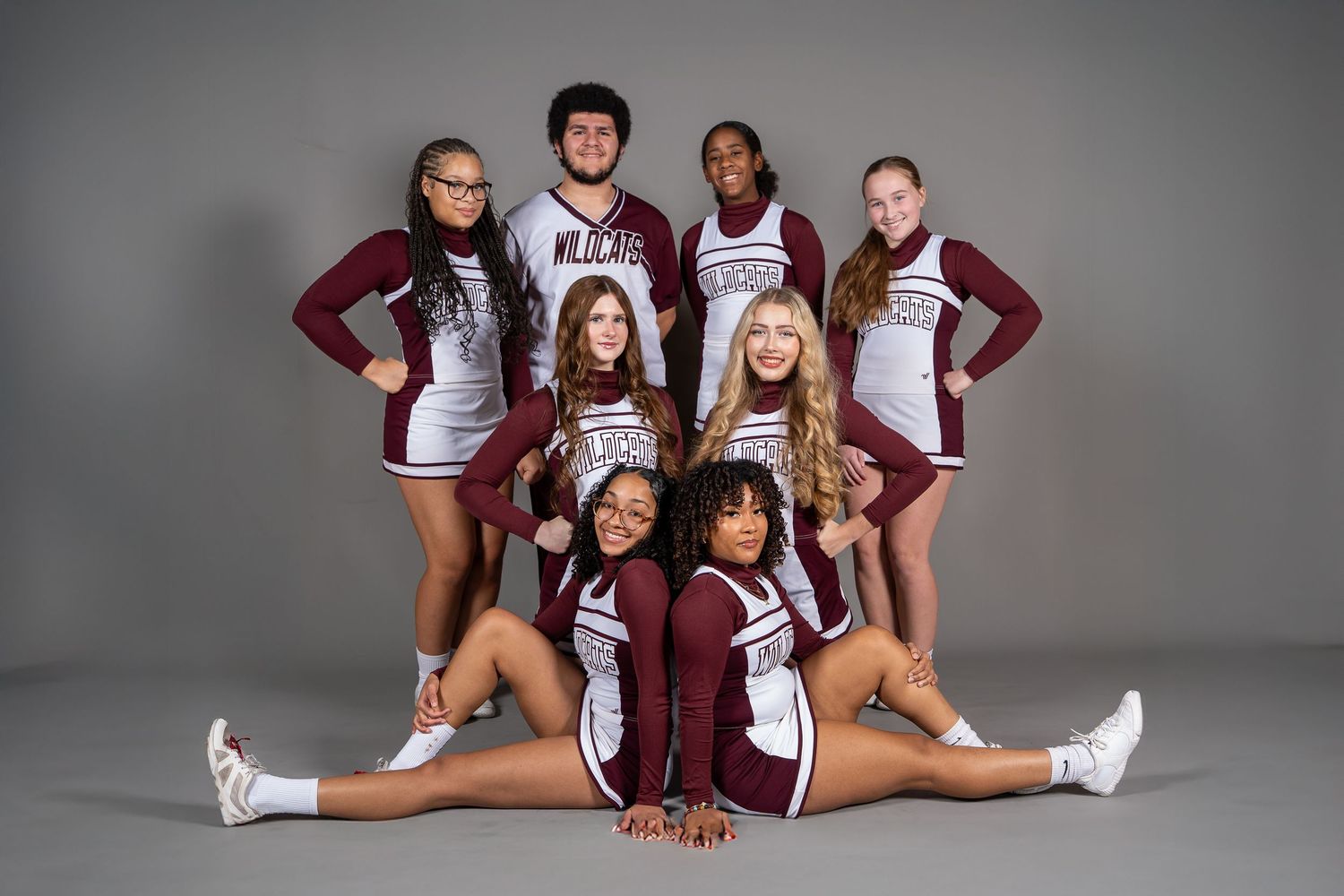 A cheerleading squad in maroon and white uniforms poses together for a team photo against a gray studio background.