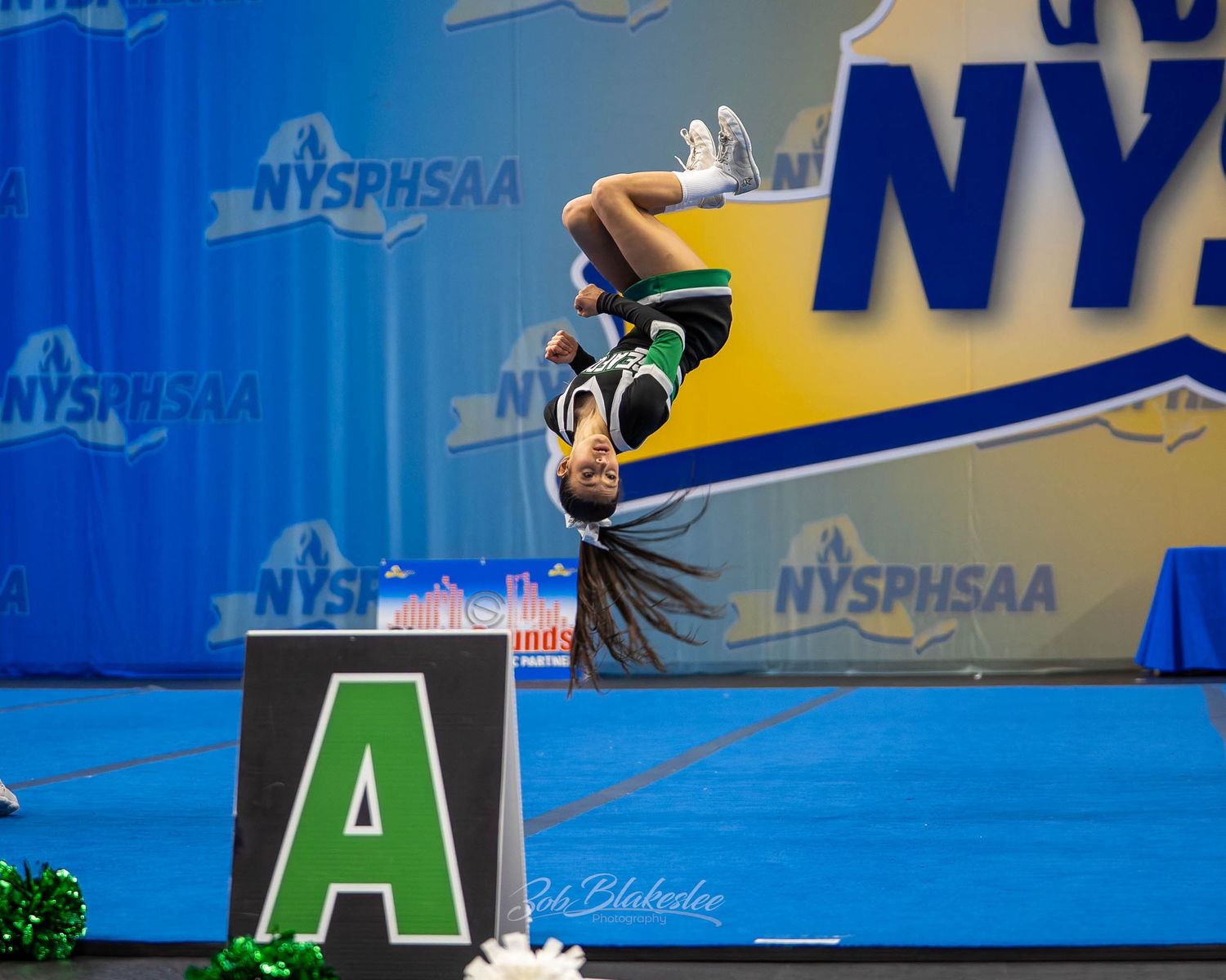 Green uniformed cheerleader performing back flip beside letter A sign at NYSPHSAA Championships.