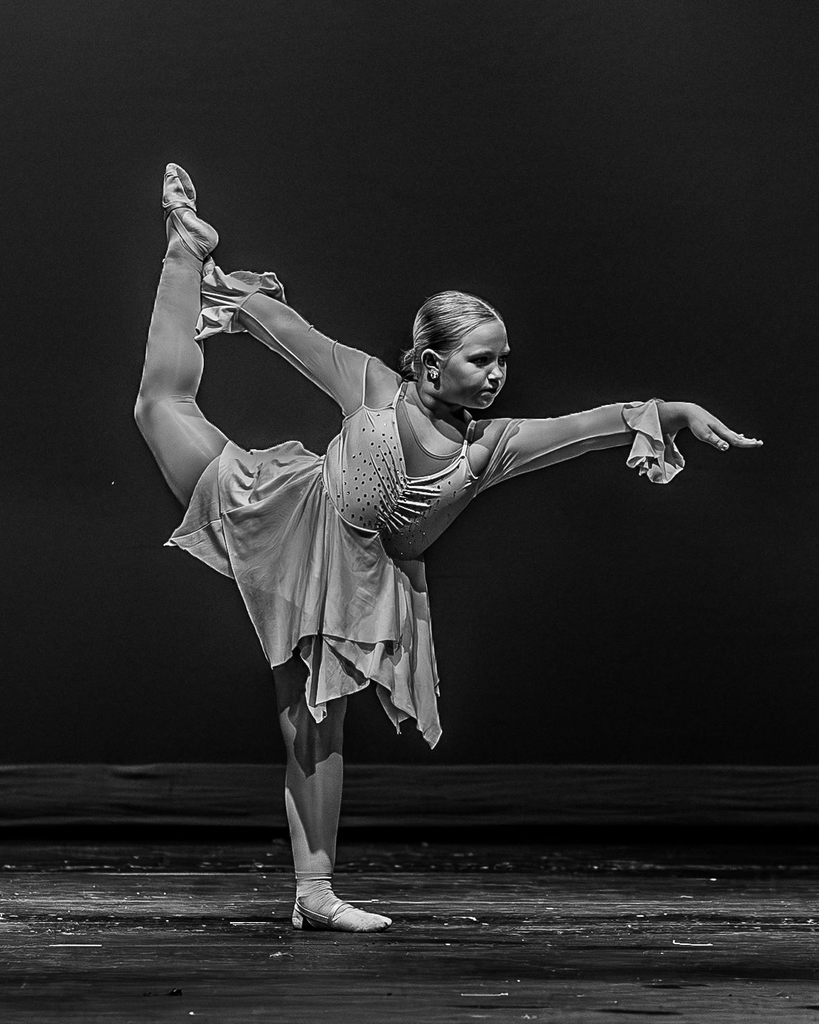 A young blonde ballet dancer performs an arabesque in a flowing costume on stage in black and white.