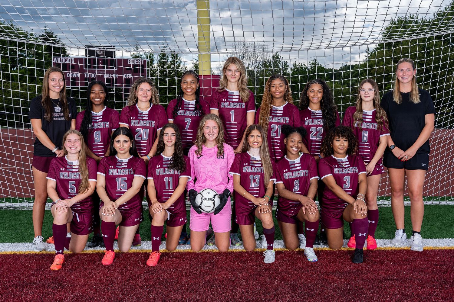Girls youth soccer team posing for official team photo in maroon uniforms on outdoor field with goal net backdrop.