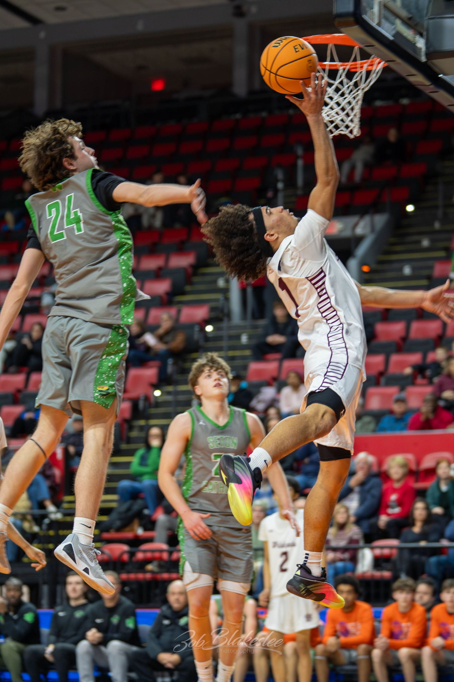 High school basketball player in white uniform drives to the hoop past defender wearing green jersey number 24.