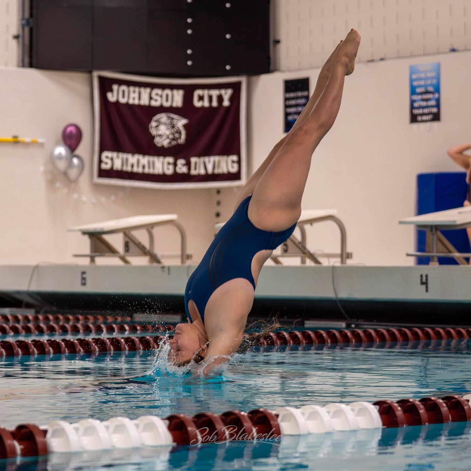 Diver performs a headfirst dive at Johnson City Swimming & Diving meet, wearing a blue swimsuit over an indoor pool.