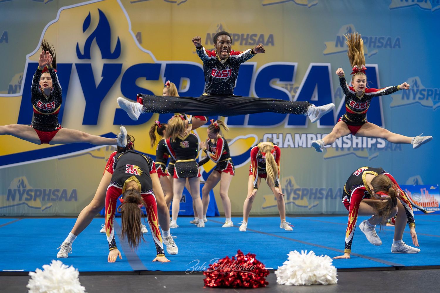 Cheerleading squad performs synchronized stunts with red pom-poms at the NYSPHSAA Championships.