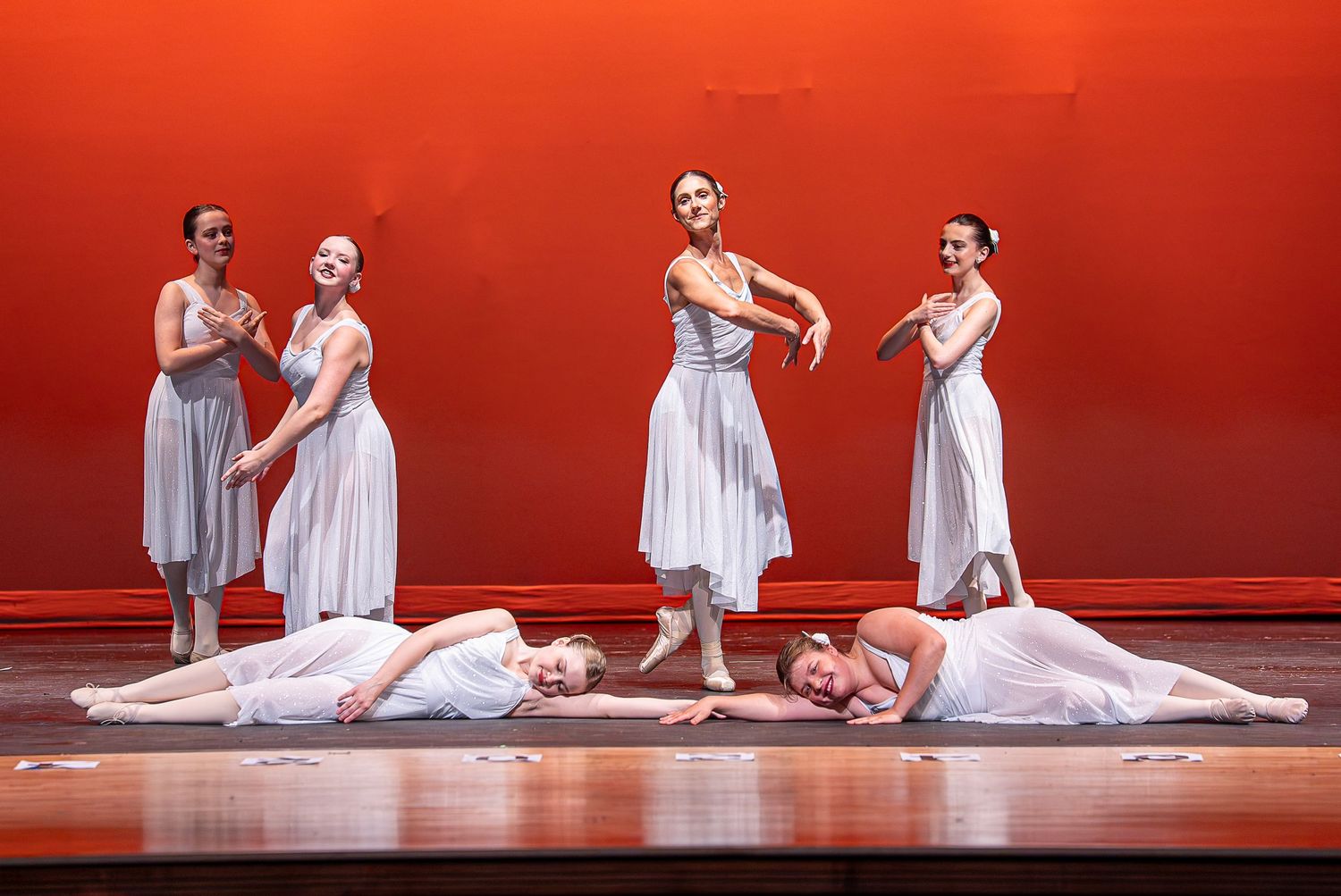 Six dancers in white dresses perform an elegant group formation on a red-lit stage.