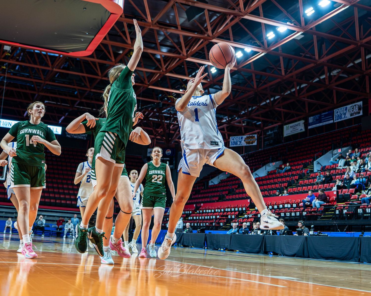Female basketball player in blue uniform attempts a shot against green-uniformed defenders in an indoor arena game.
