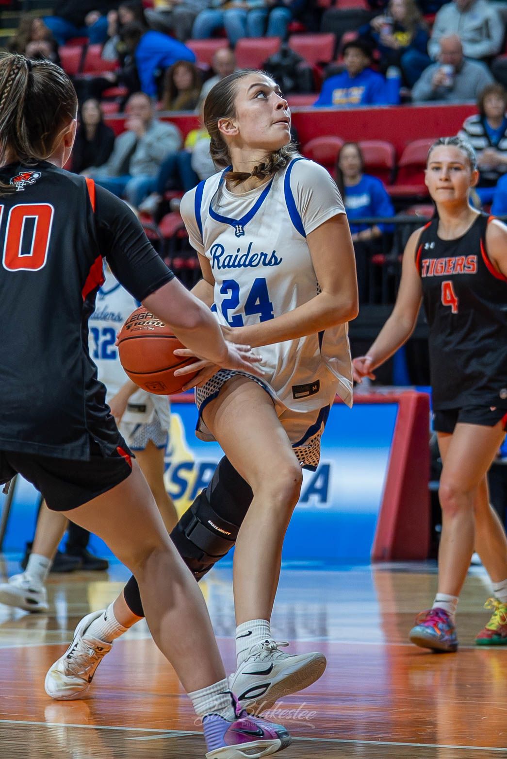 Female basketball player #24 in blue Raiders jersey drives to the basket during a competitive high school game.