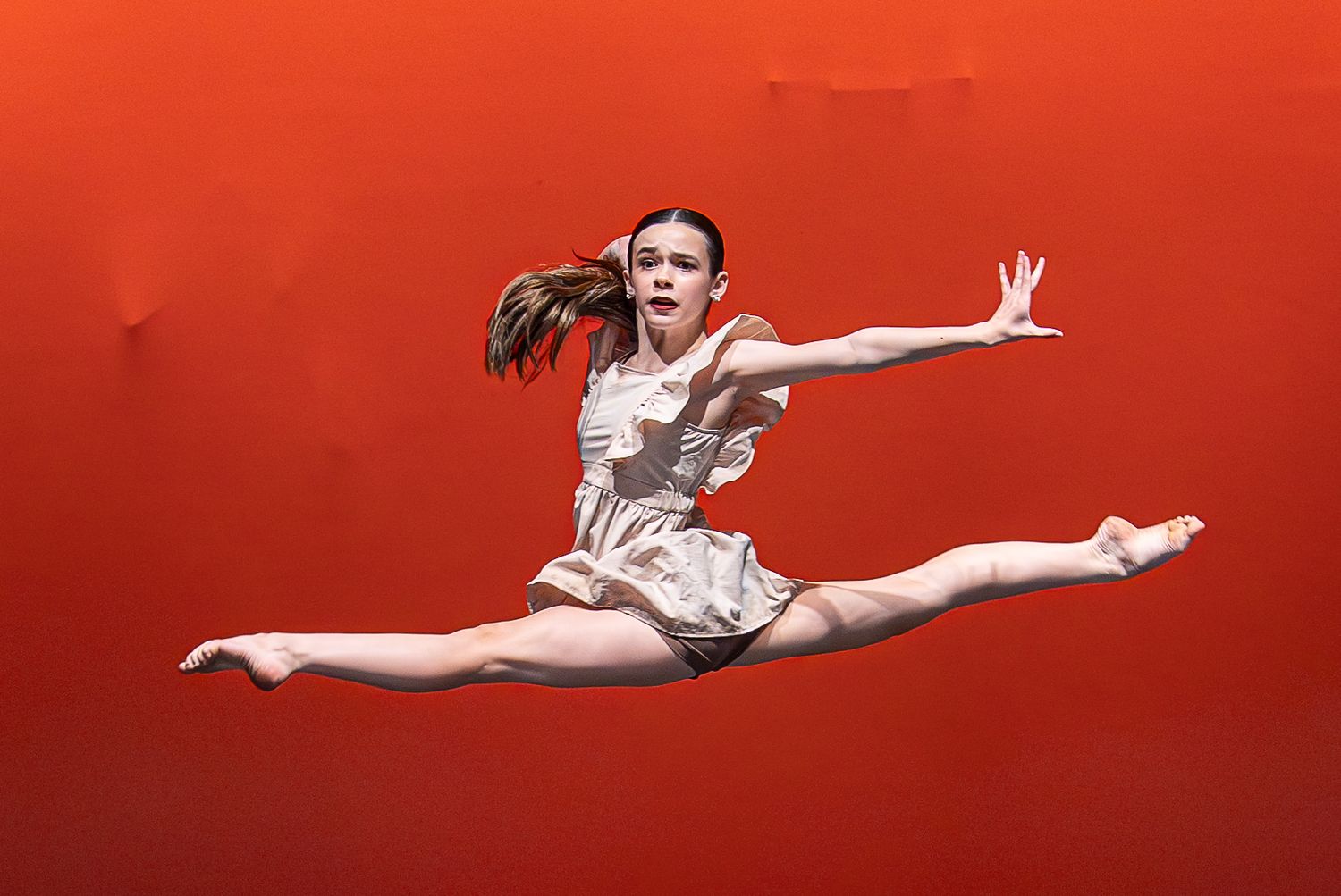 A young dancer performs a perfect split leap against a bold red background on stage.