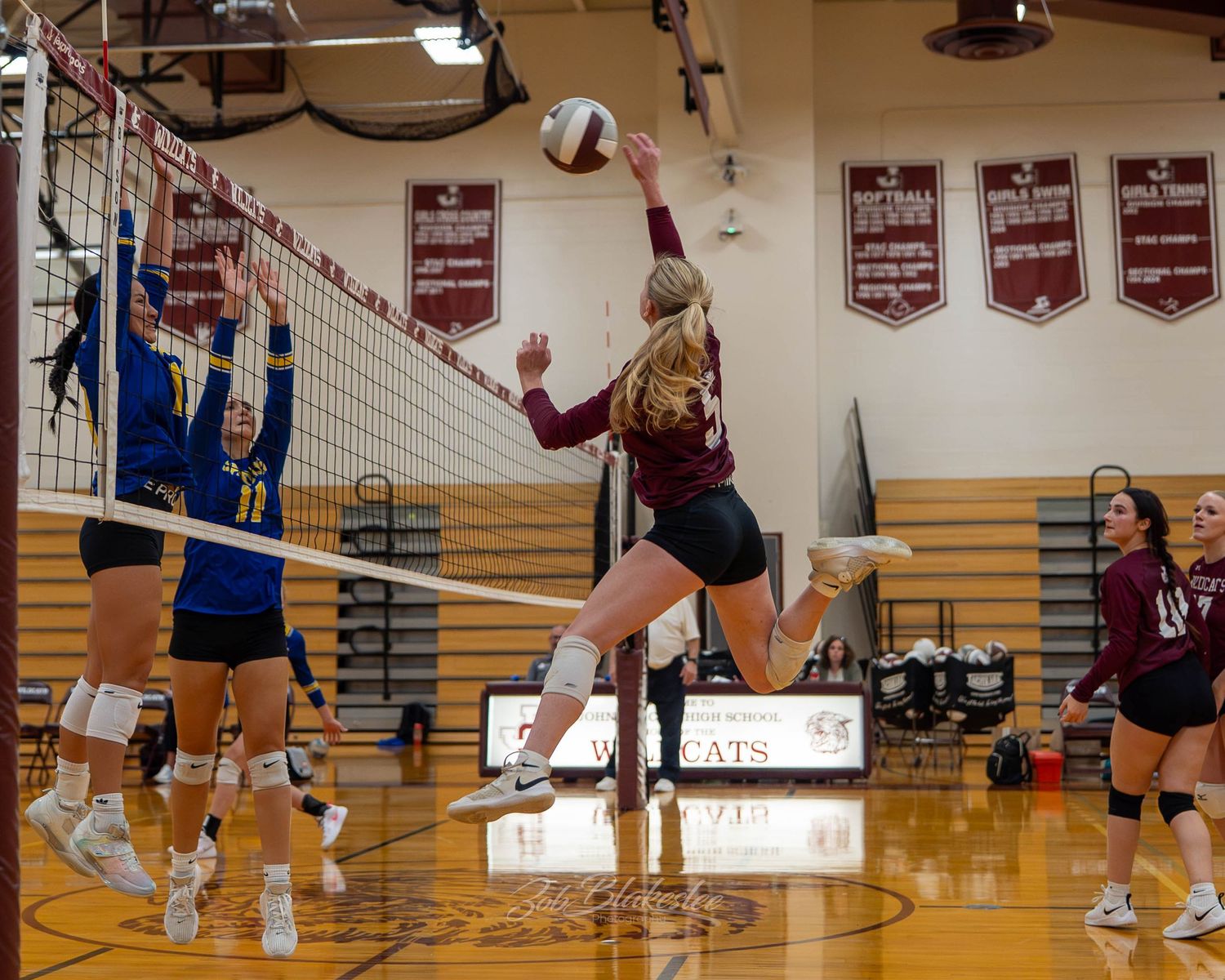 A volleyball player leaps to spike the ball over the net during an intense high school gym match.