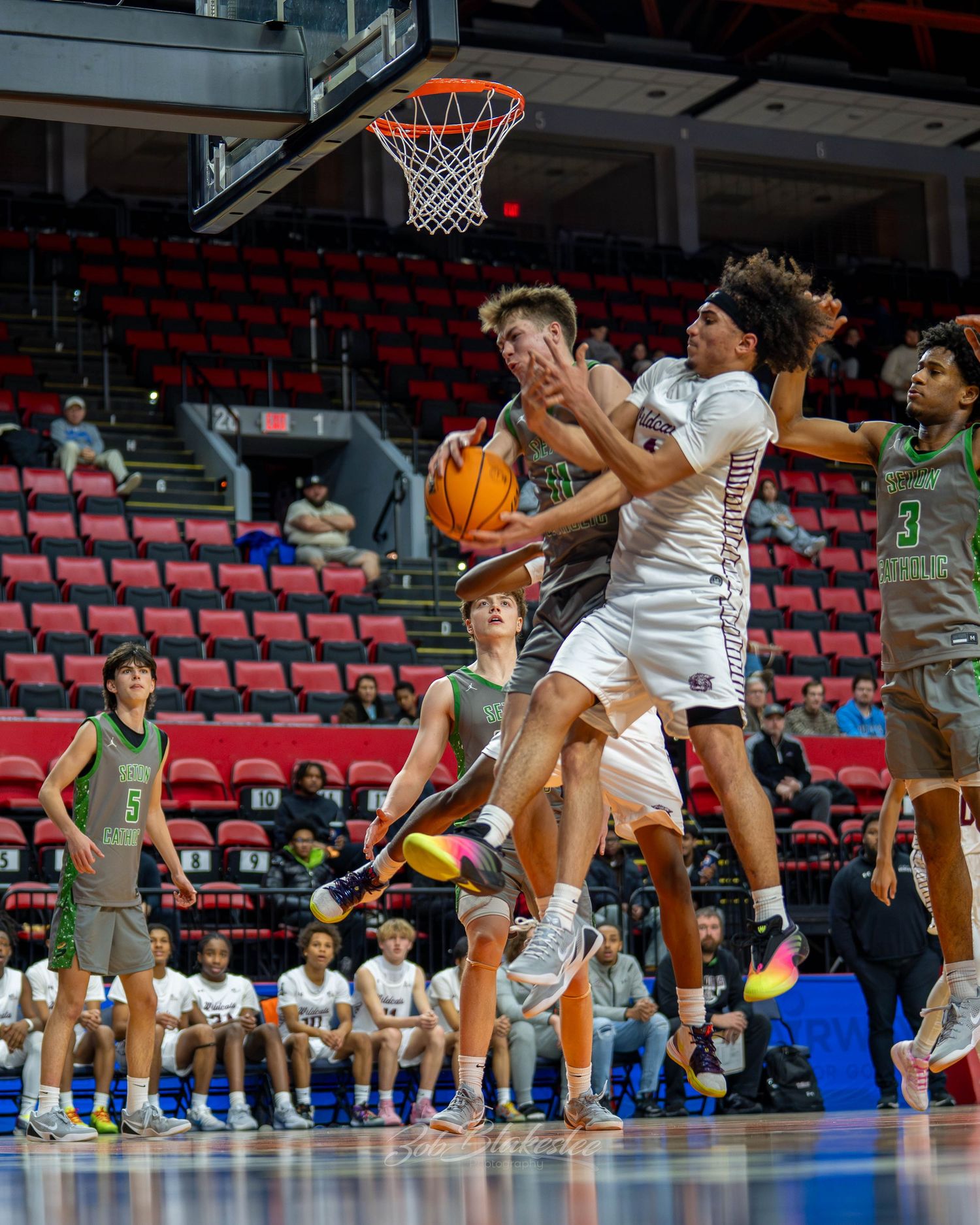 Male basketball players compete for a rebound during an indoor arena basketball game with empty red seats.