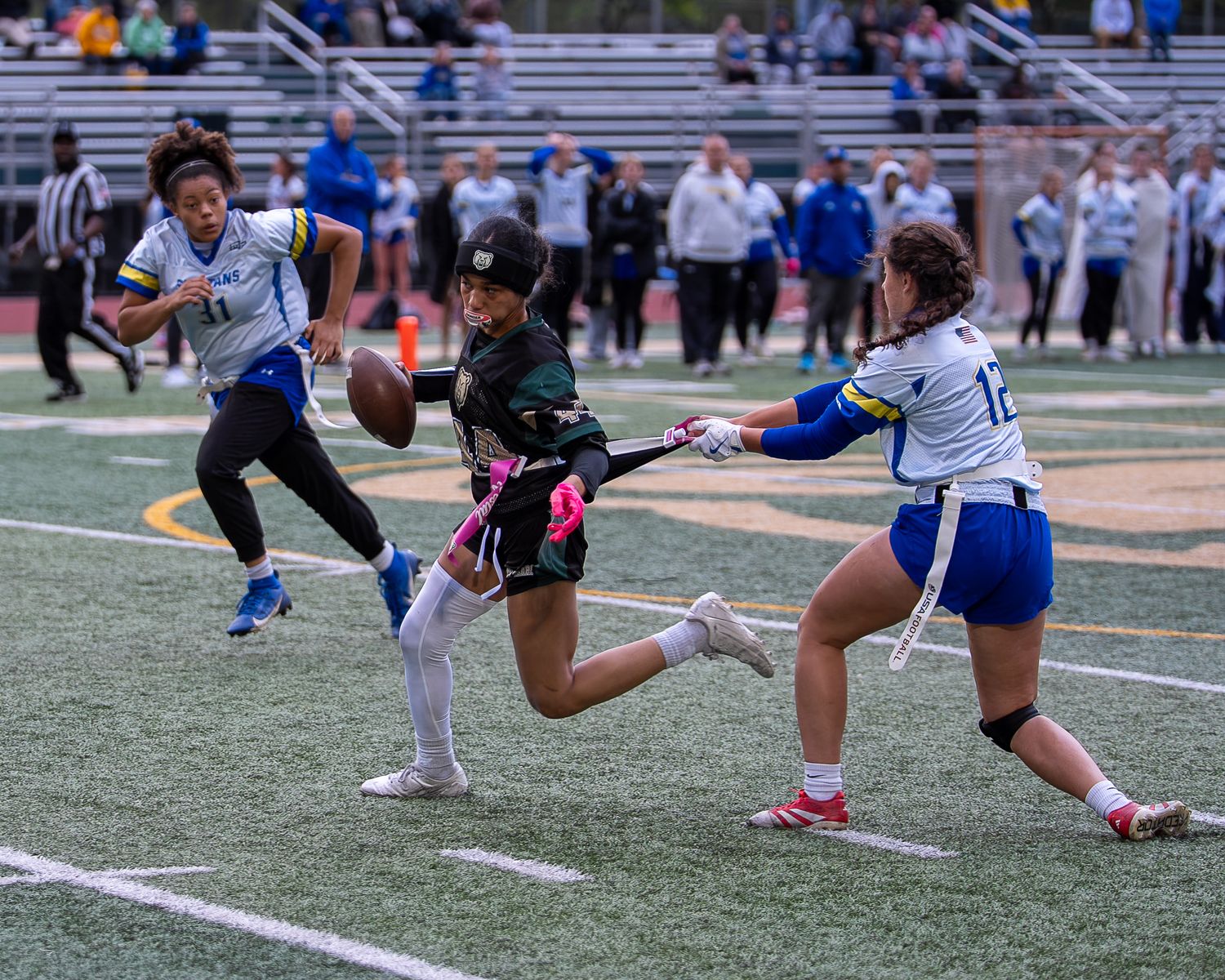 Female athletes compete in a flag football game, with one player catching the ball while defenders attempt to pull flags.