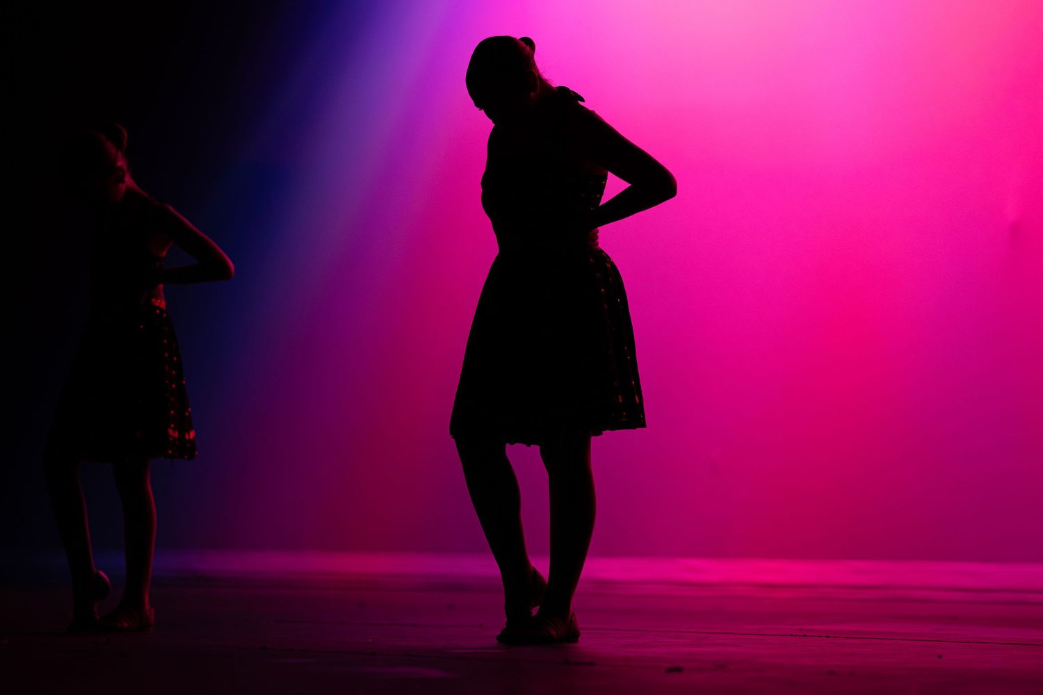 Two silhouetted dancers perform against vibrant pink and blue stage lighting in the dark.