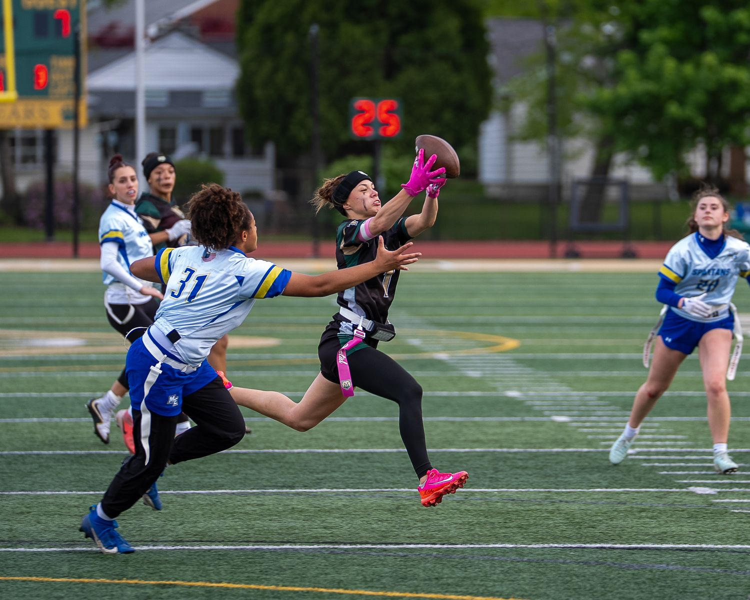 Women playing flag football, player in black catching pink ball while defender in blue jersey number 31 attempts block.