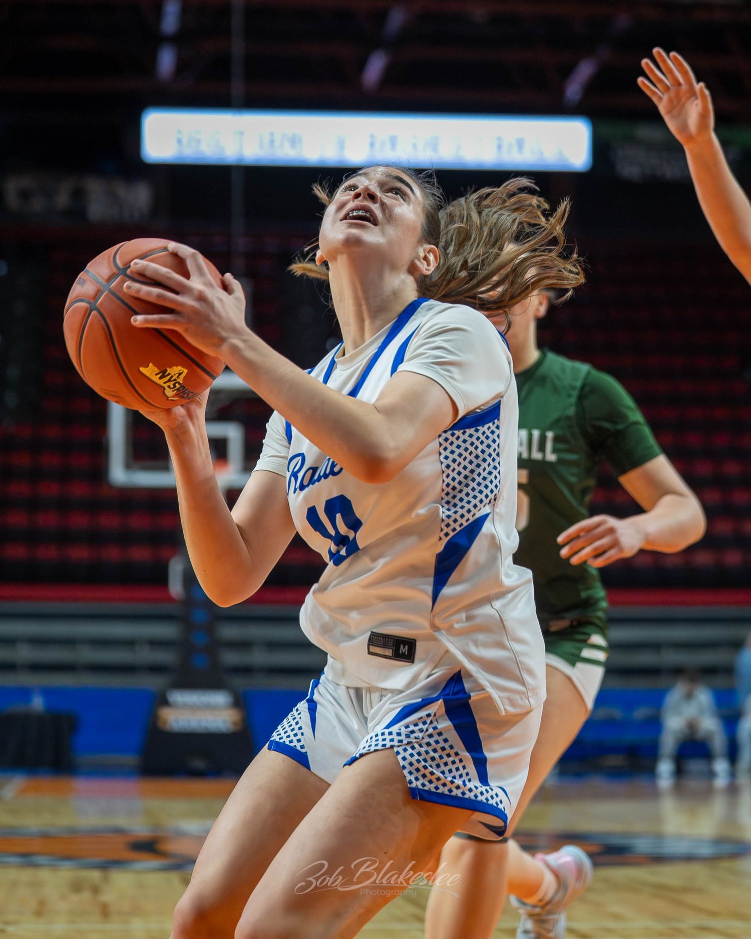 Female basketball player in blue uniform drives to the basket during a competitive high school game.