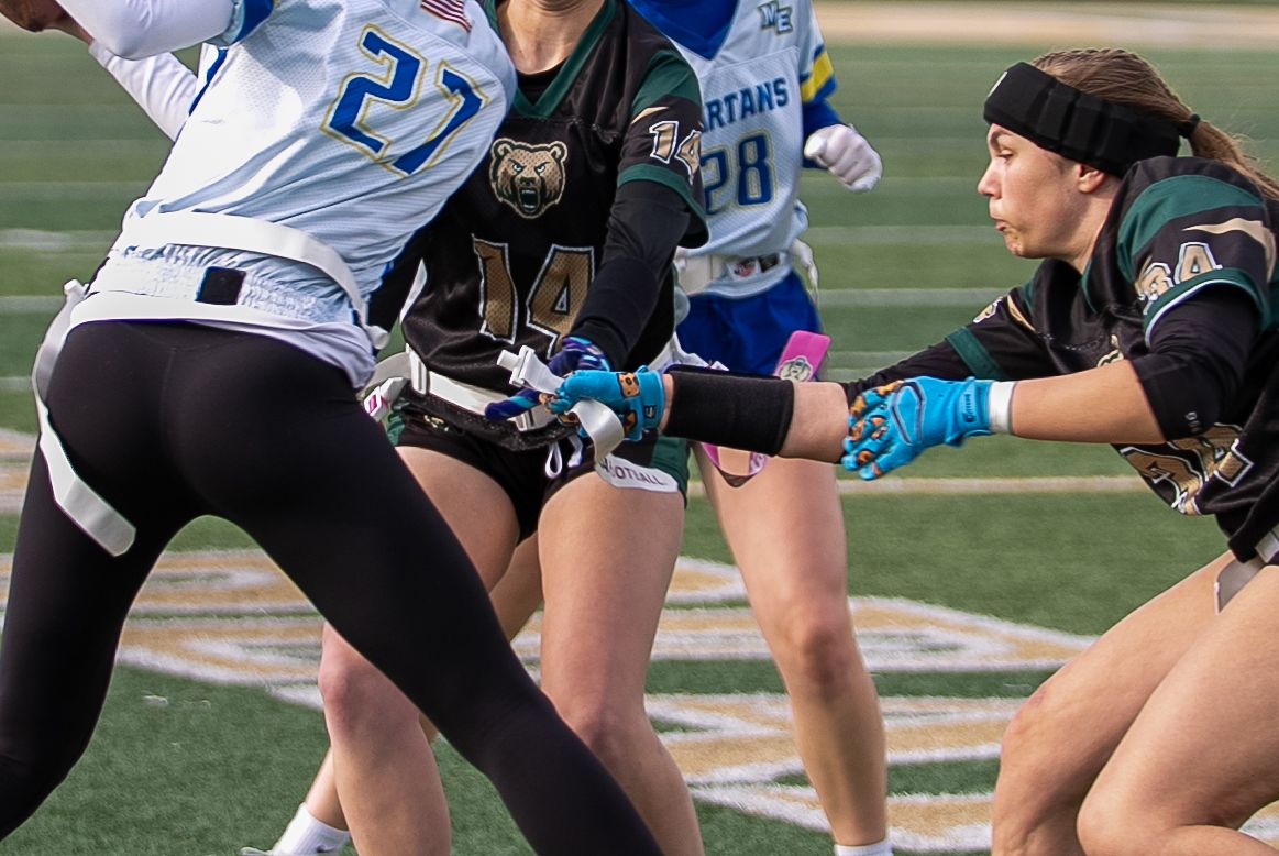 Women playing flag football on a field, with players in green and white uniforms competing during a game.