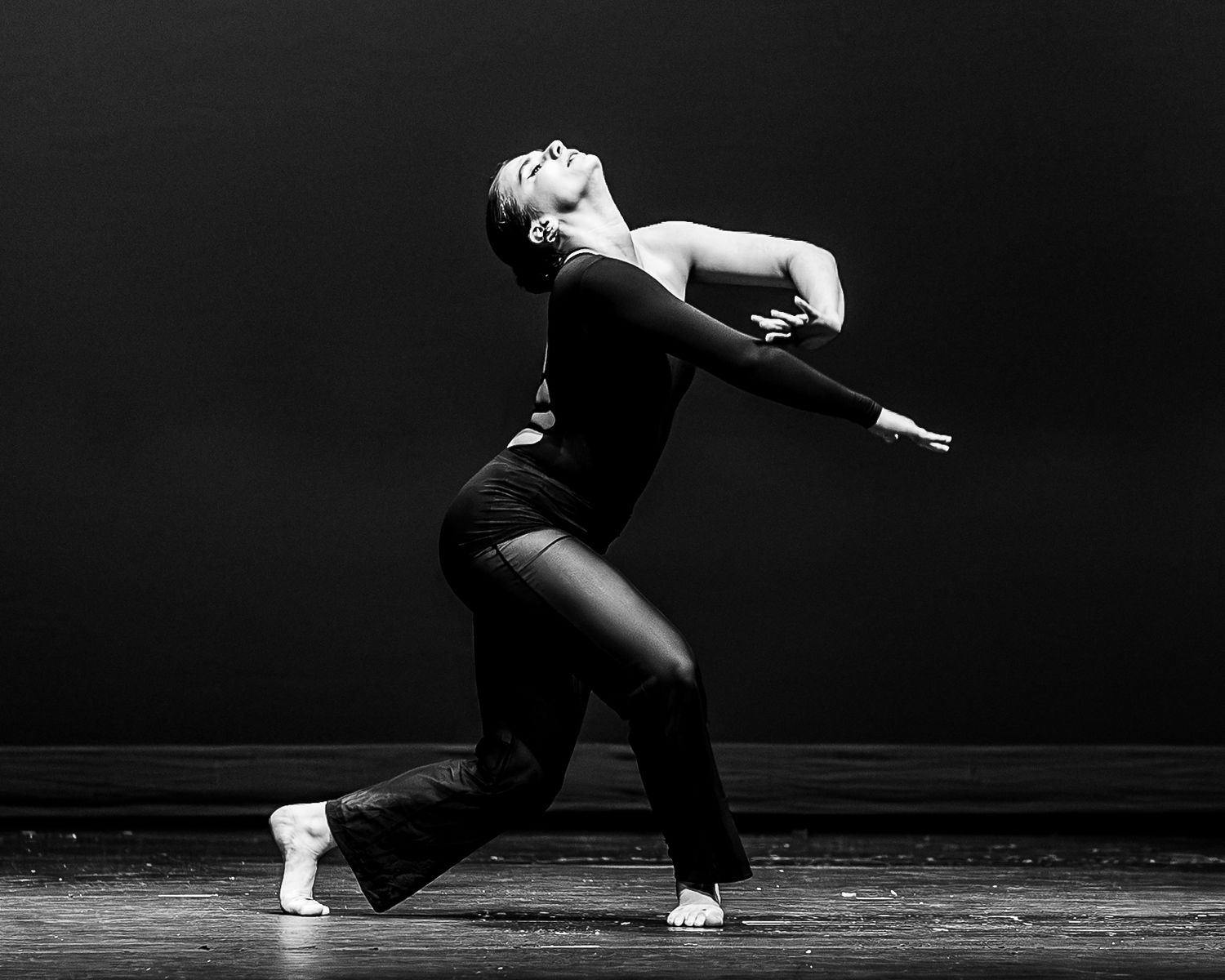 A solo contemporary dancer arches dramatically in black attire on a dark stage in black and white.