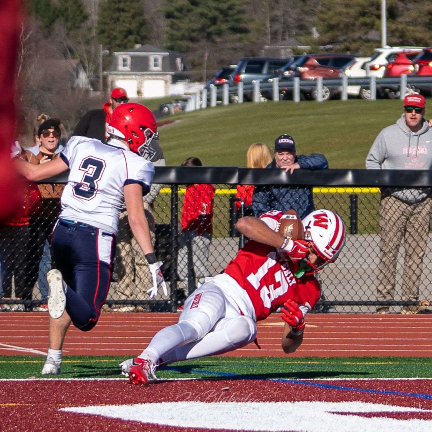 Football player makes diving catch in end zone during high school playoff game on sunny day.