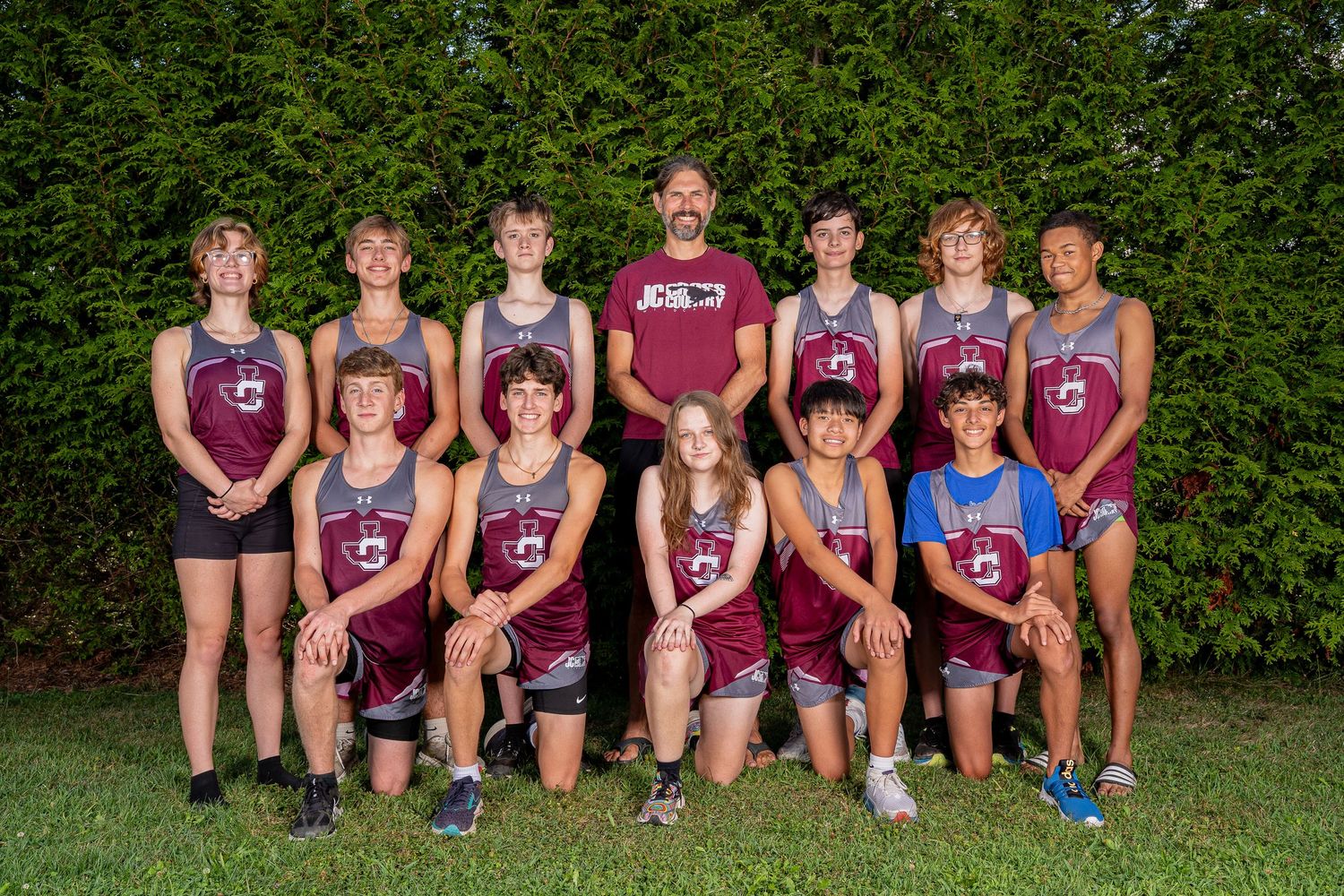 High school cross country team poses outdoors in maroon uniforms with coach, two rows, green hedge background.