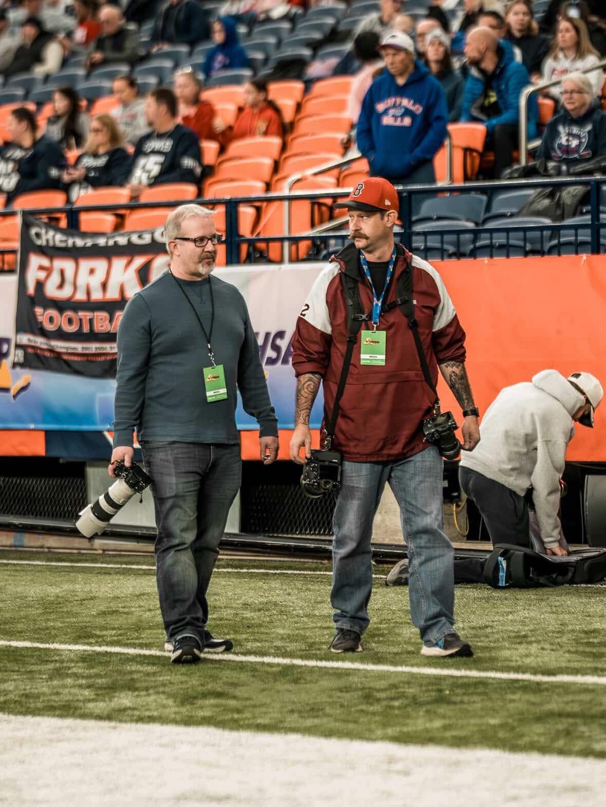 Two photographers with cameras and green press passes walk on a football field during an indoor stadium game.