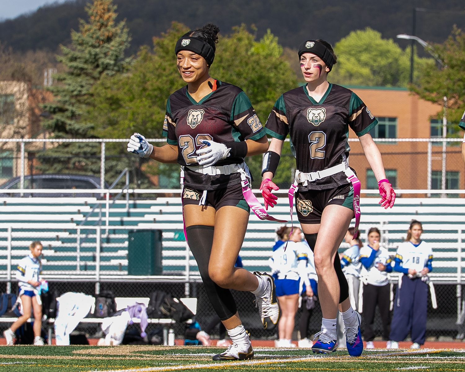 Two female flag football players in dark uniforms running on a field during a sunny outdoor game.