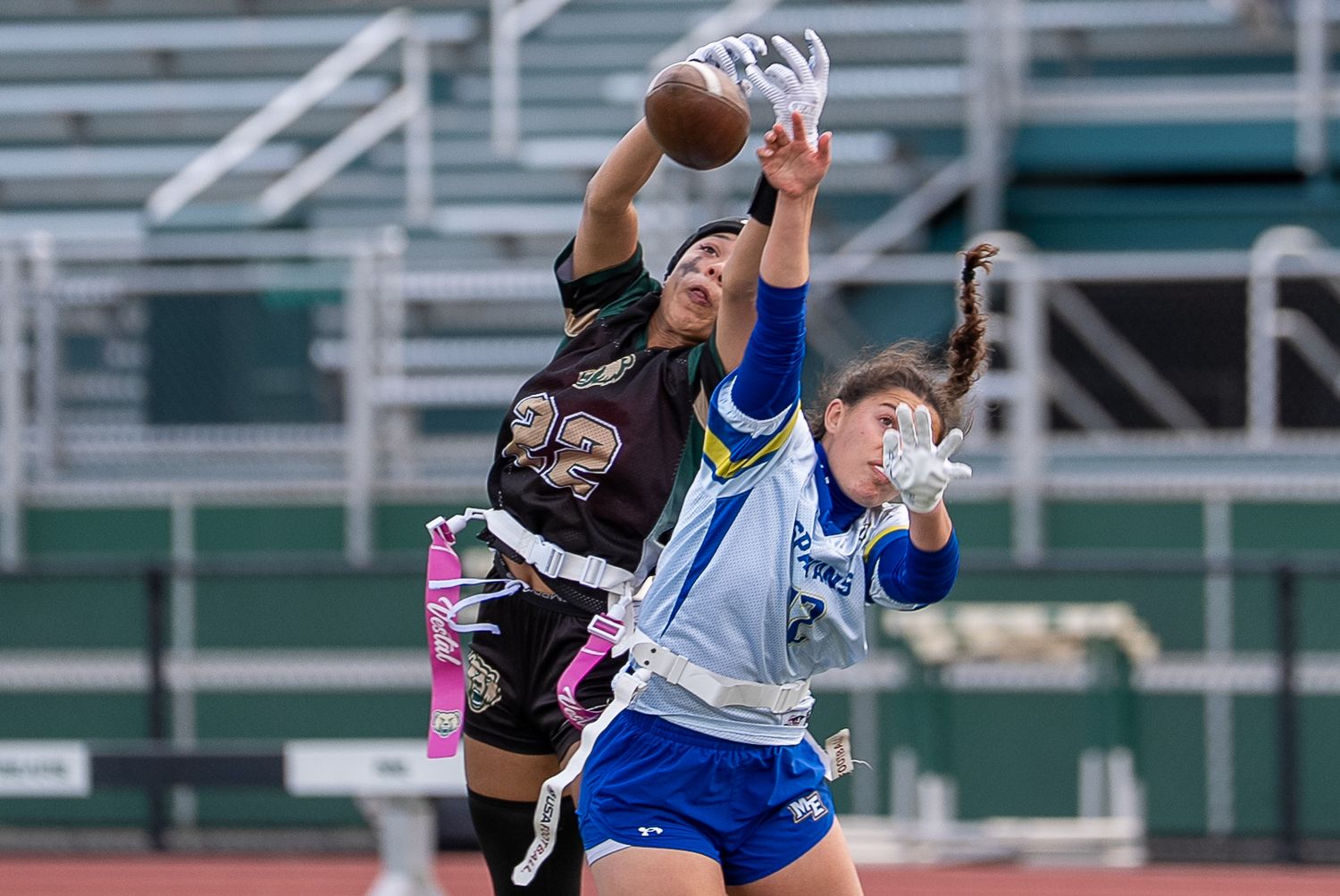 Two female athletes compete for a football catch during an intense flag football game on a green field.
