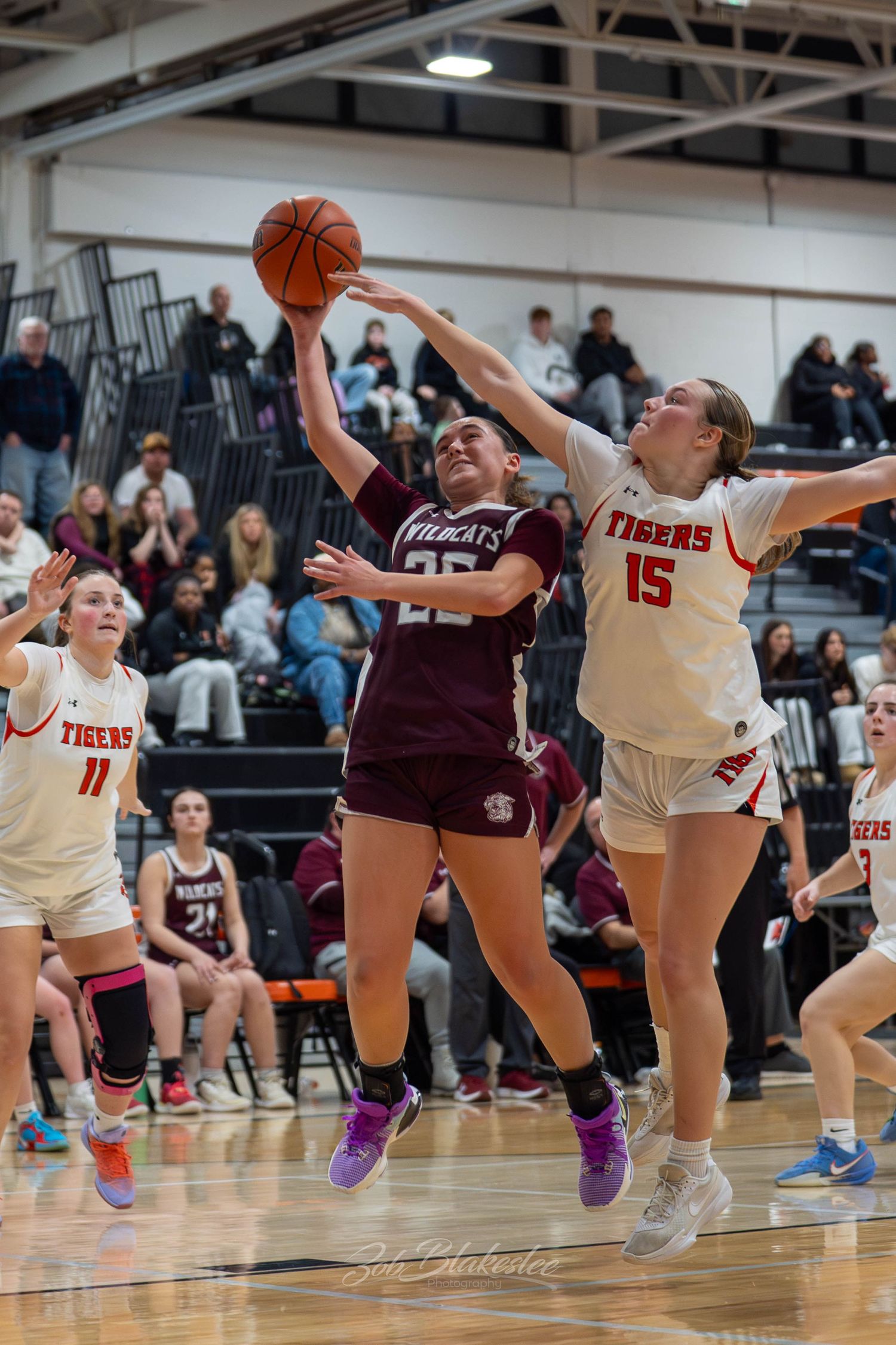 Female basketball players compete in a high school game, with a Wildcats player driving to the basket against a Tigers defender.