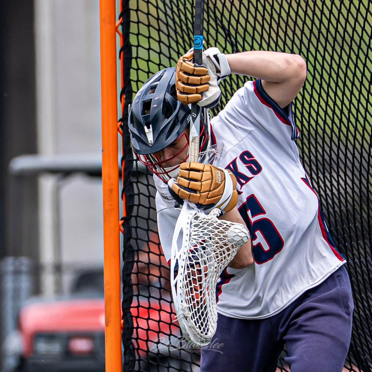 A lacrosse goalie in white jersey cradles the ball in the net during an outdoor game.