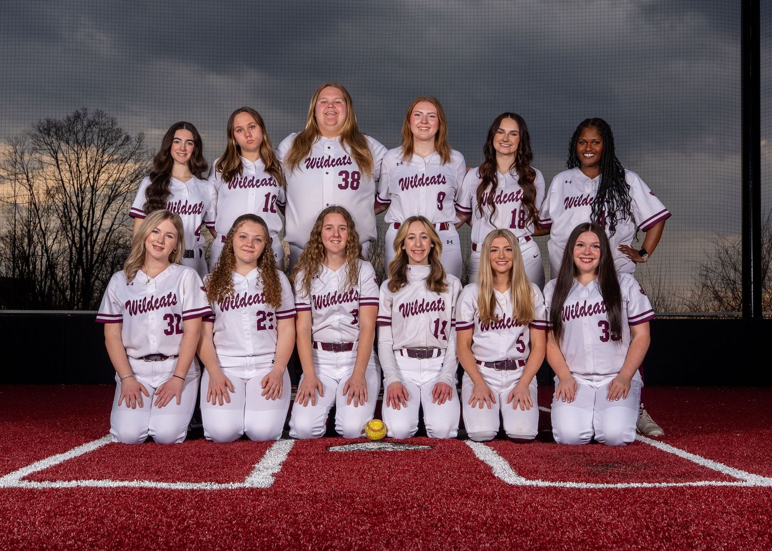 Wildcats softball team poses in white uniforms on a red field under dramatic cloudy skies for their official team photo.