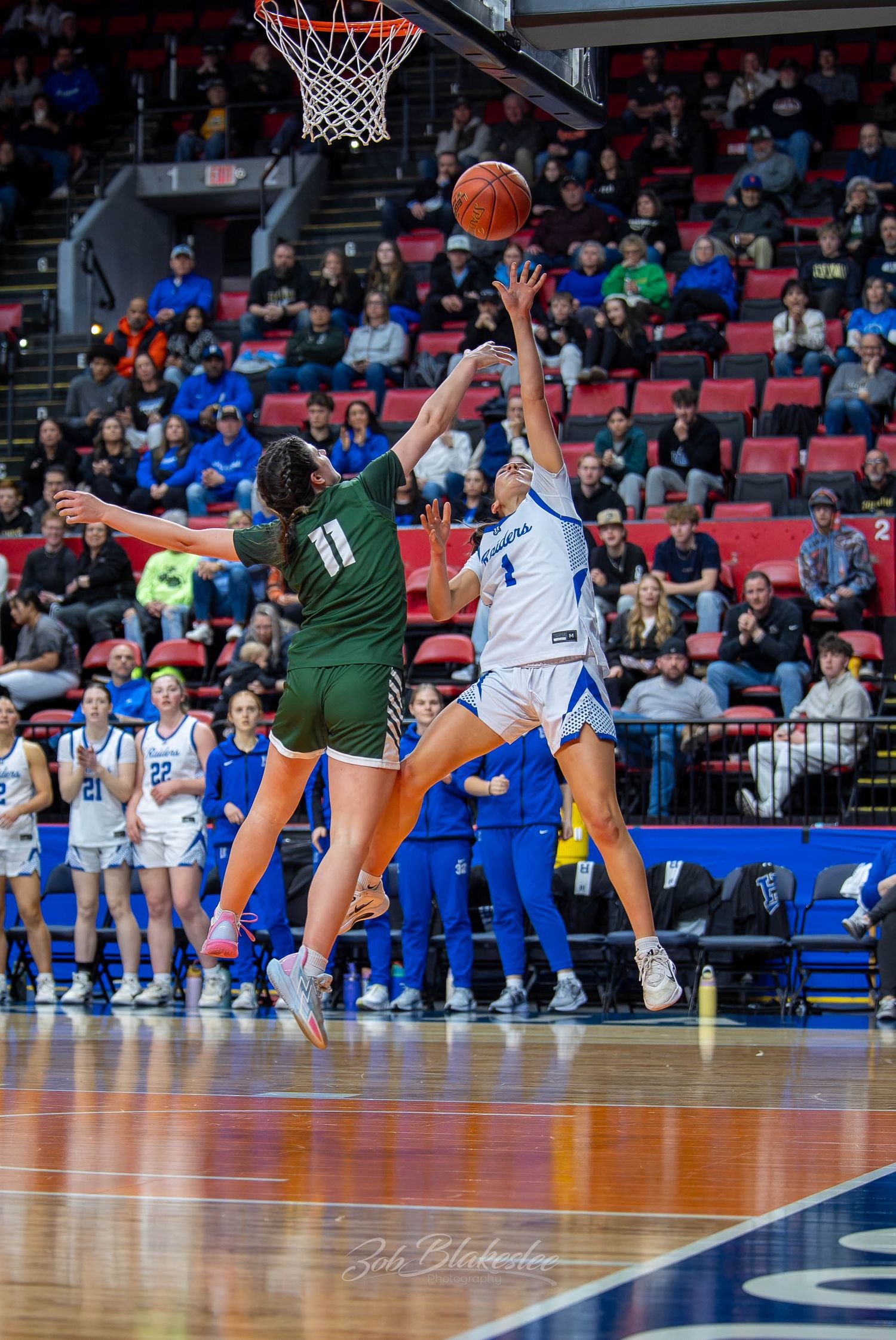 Two female basketball players compete for a shot near the hoop during a packed arena game, wearing green and blue uniforms.