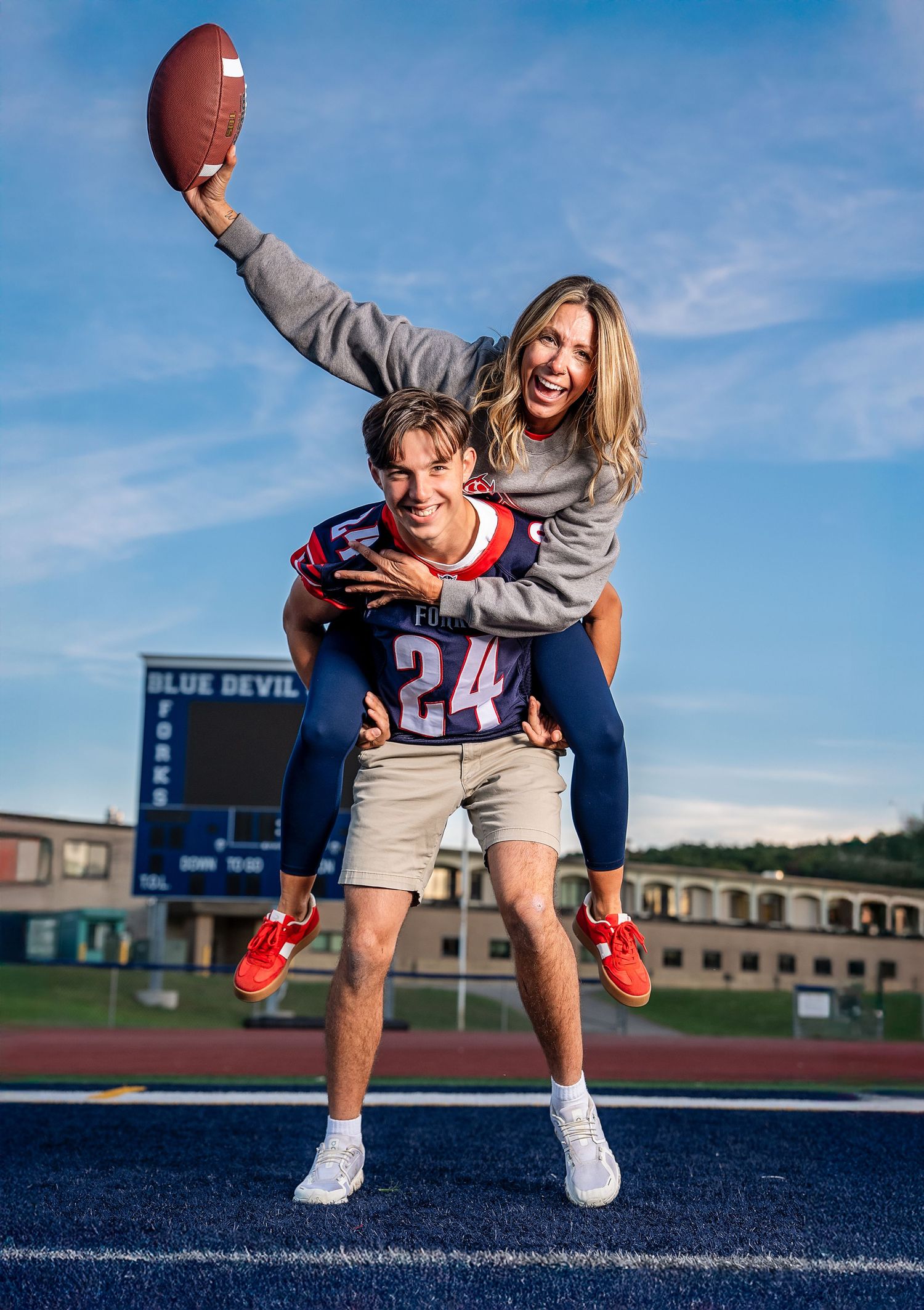 Young man in football jersey #24 gives a woman a piggyback ride on a blue turf field as she holds a football.