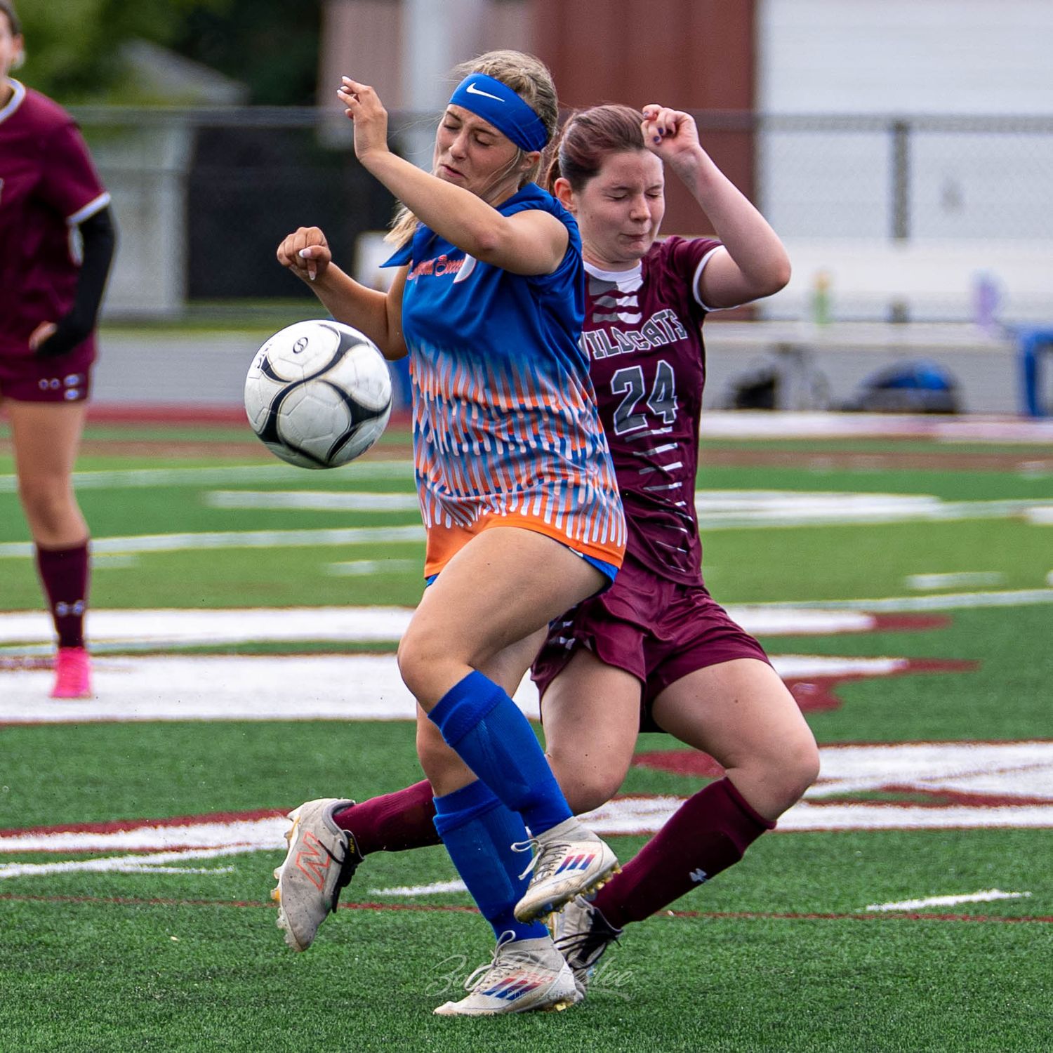 Two female soccer players compete for the ball during a match, one in blue and orange, the other in maroon uniform.