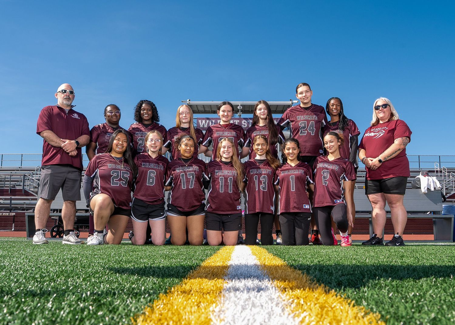 Girls flag football team poses on field in maroon jerseys with coaches on a sunny day.