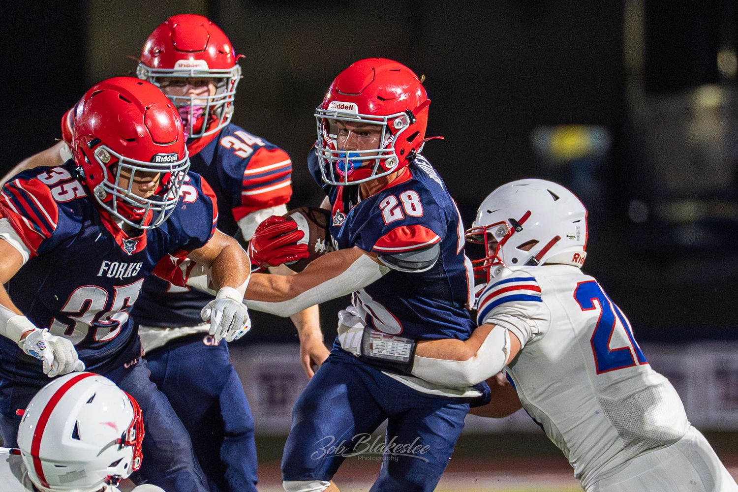 High school football players in red and blue uniforms compete during an intense night game under stadium lights.
