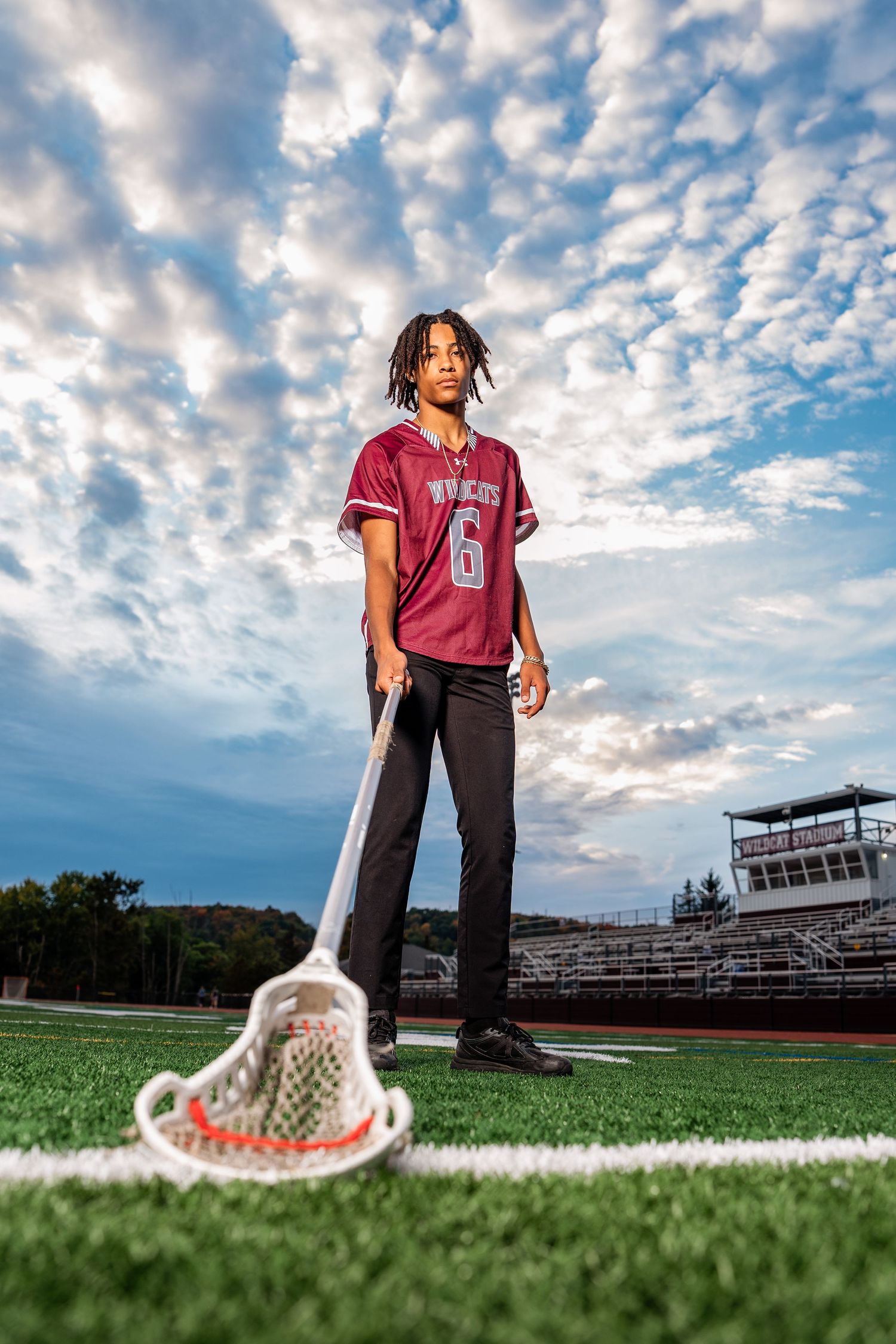 A lacrosse player in maroon jersey number 6 poses with stick on turf field under dramatic cloudy sky.