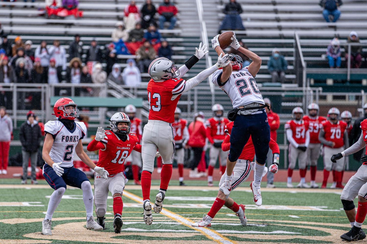 High school football players compete for a pass during an outdoor game on a green turf field in cold weather.