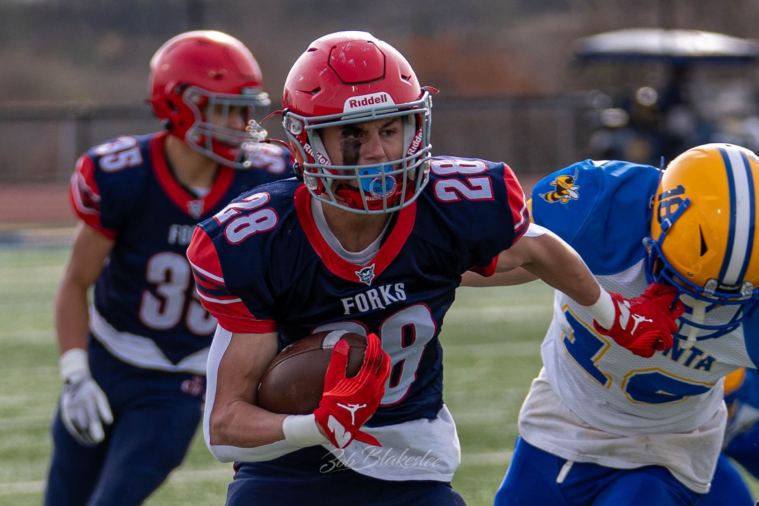 High school football player wearing number 28 runs with the ball during a daytime outdoor game.