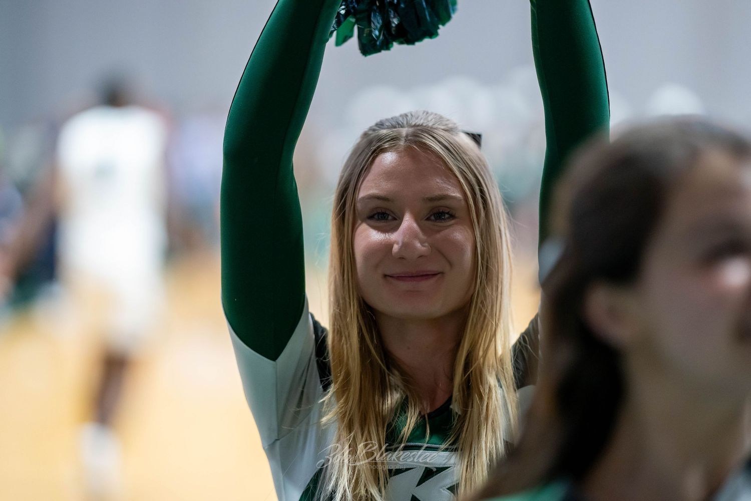 A smiling female cheerleader in green uniform raises pom-poms overhead during a basketball game.
