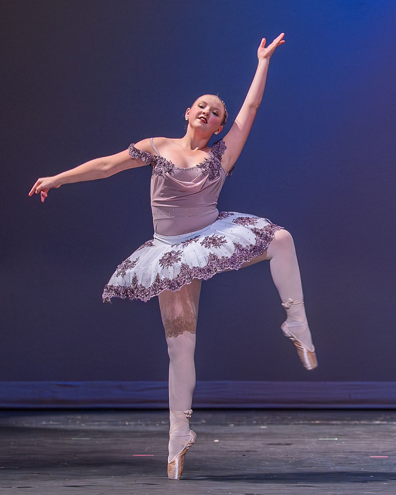 A ballerina en pointe in a white tutu with floral details performs on a softly lit stage.