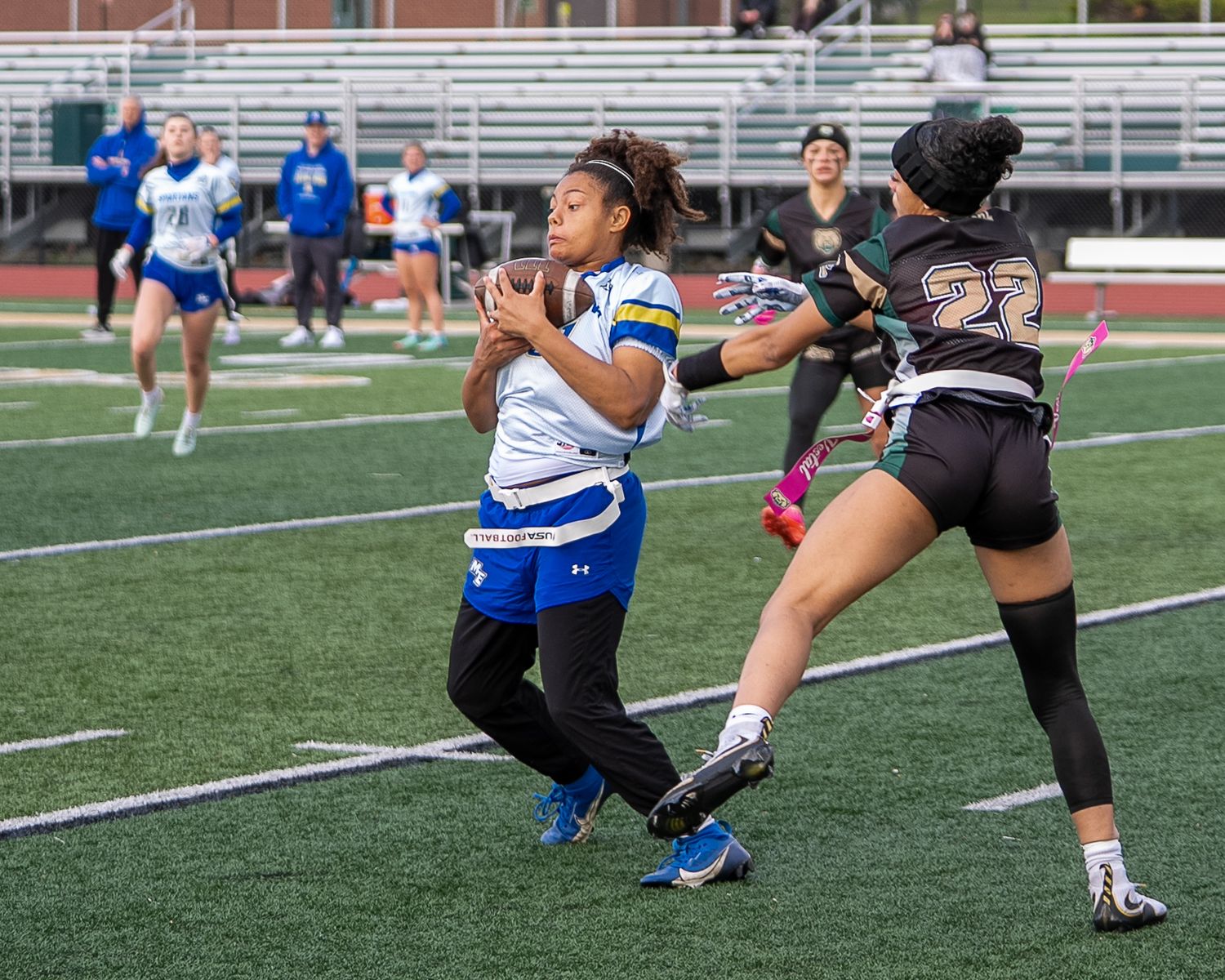 Two female flag football players compete on a turf field, one attempting to grab the flag from the other.