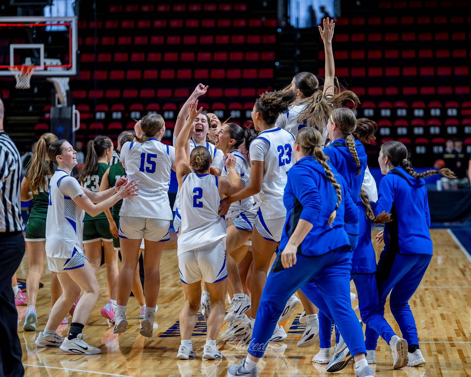 Girls basketball team celebrates victory on court, players in blue and white uniforms cheering together in arena.