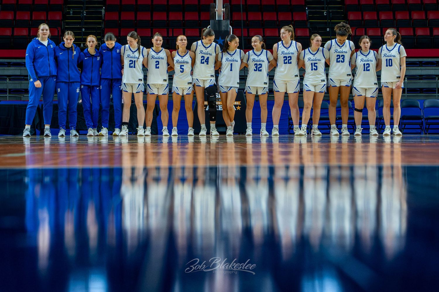 Women's basketball team poses in blue and white uniforms on a reflective hardwood court inside an indoor arena.